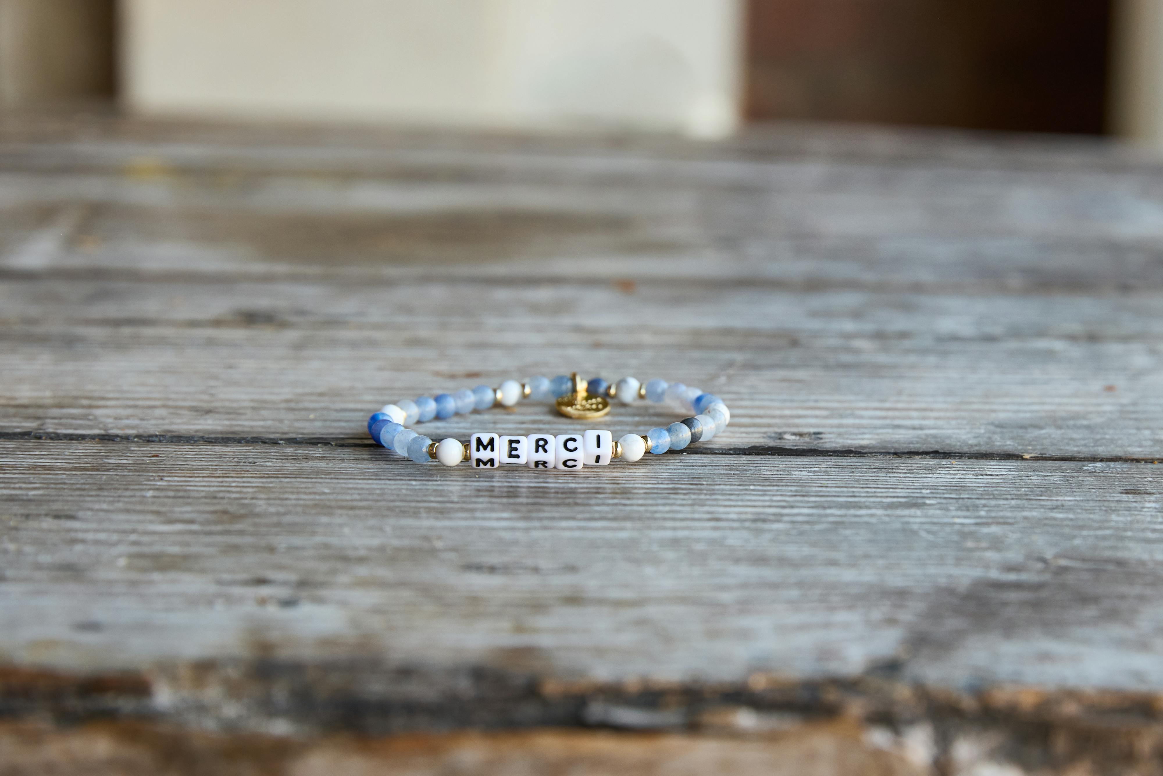 a blue beaded bracelet that spells "merci" resting on a rustic wooden table