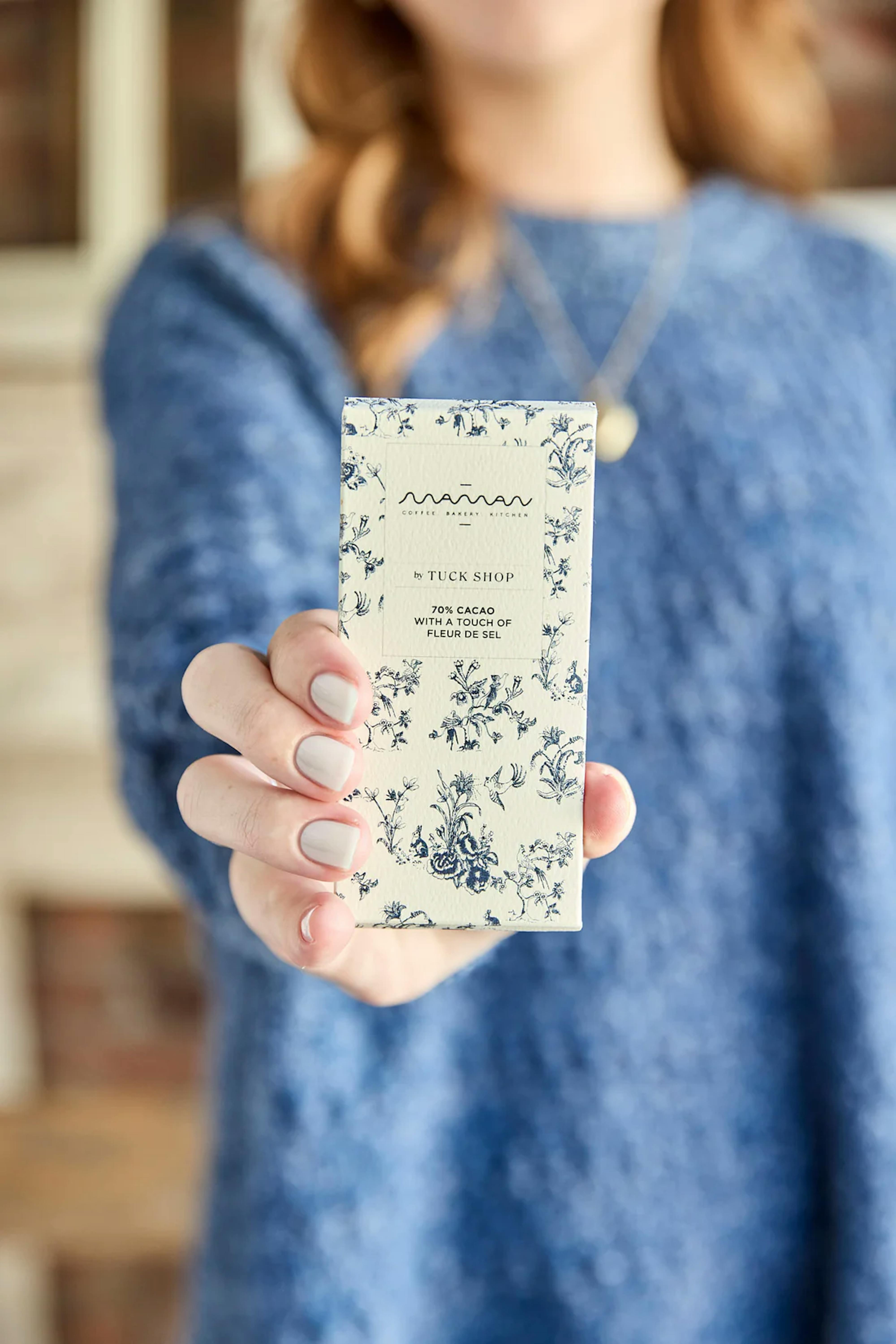 person in a blue sweater holding a maman x tuck shop artisanal chocolate bar with blue floral packaging, featuring 70% cacao and a touch of fleur de sel
