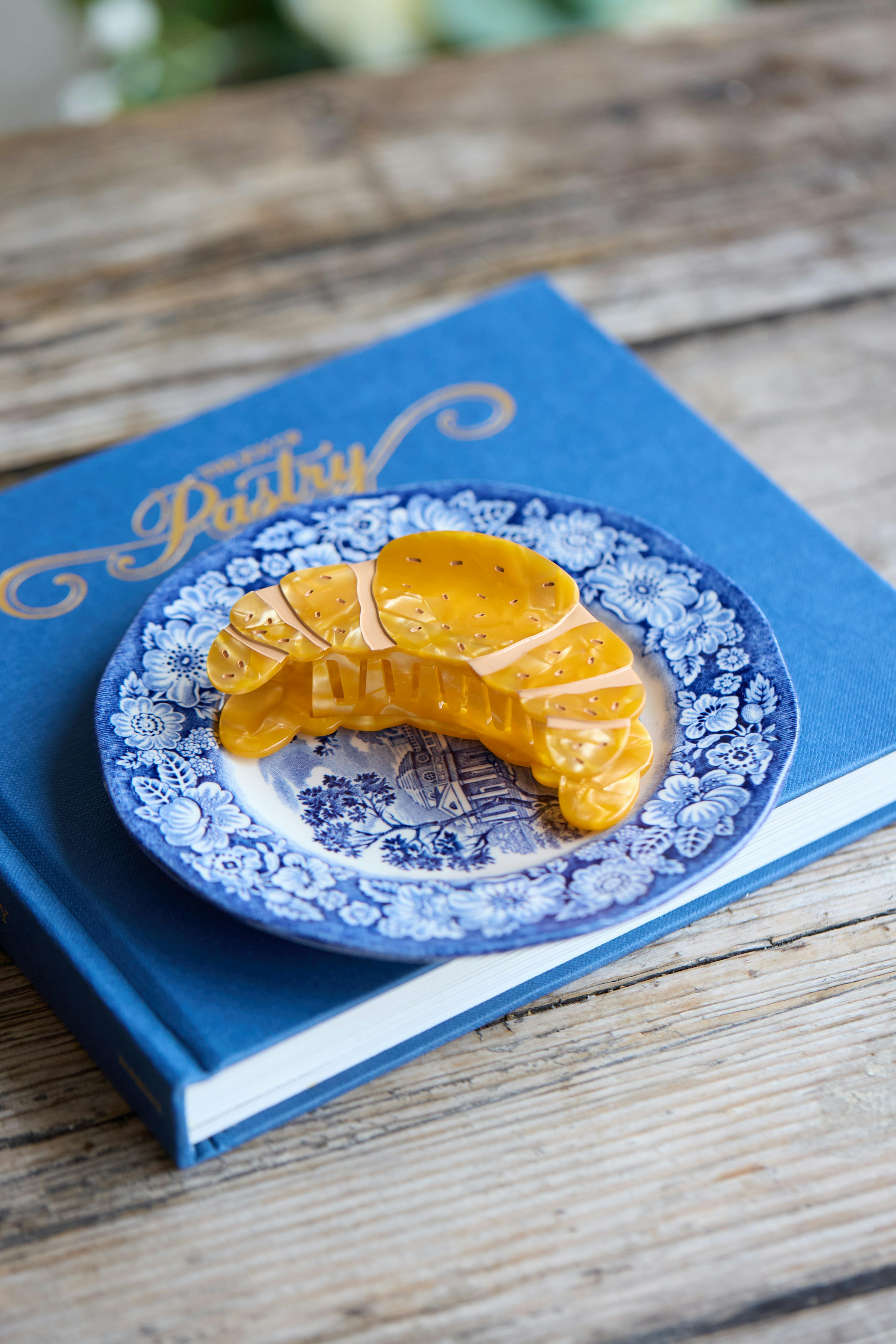 yellow croissant-shaped hair clip resting on a blue and white floral plate, placed on top of a blue pastry cookbook on a rustic wooden table