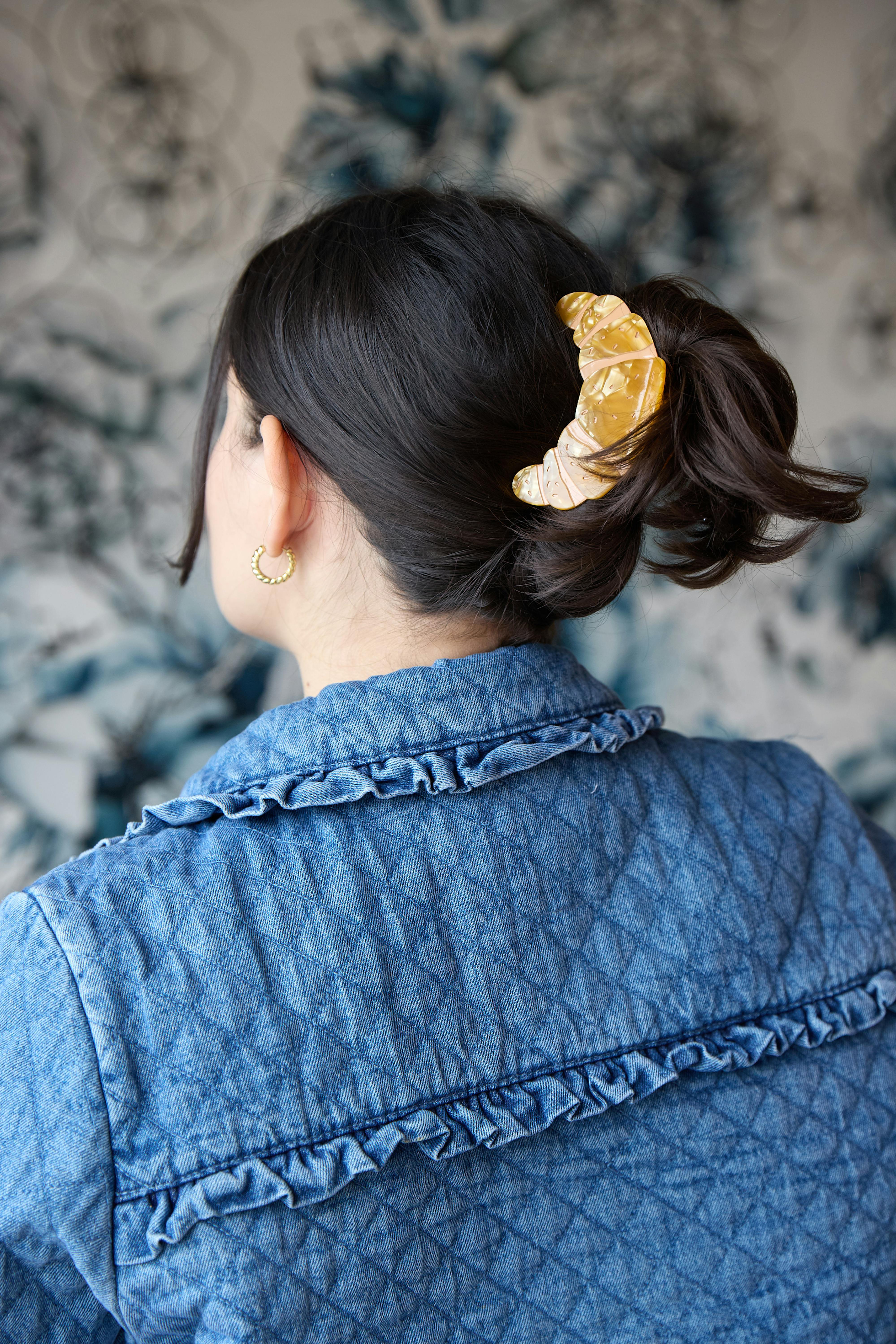 woman wearing a denim jacket with a yellow croissant-shaped hair clip securing her dark hair in an updo, against a blue and white floral background
