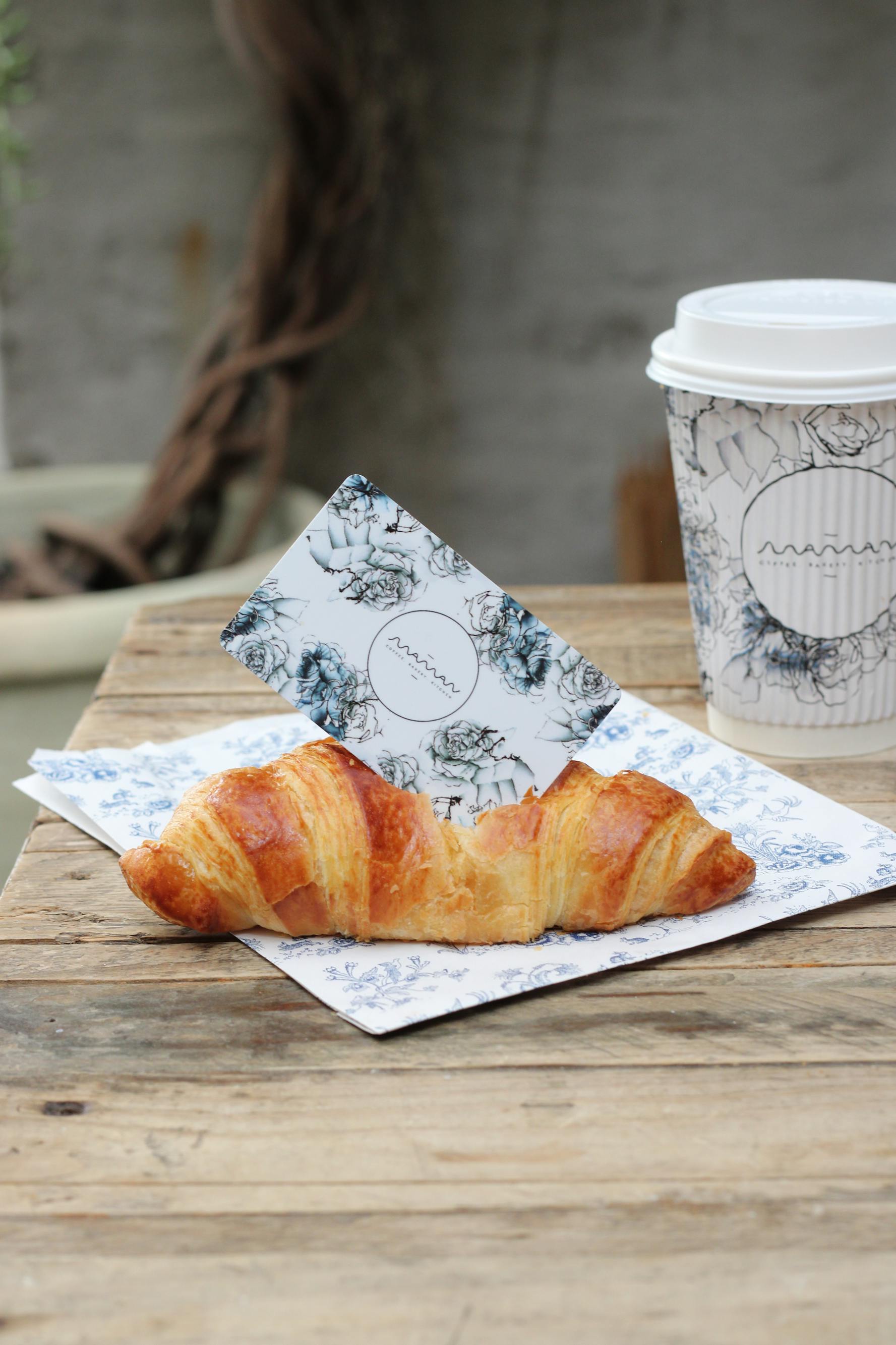 maman gift card tucked into a buttery croissant, placed on a blue floral napkin with a maman coffee cup in the background on a rustic wooden table