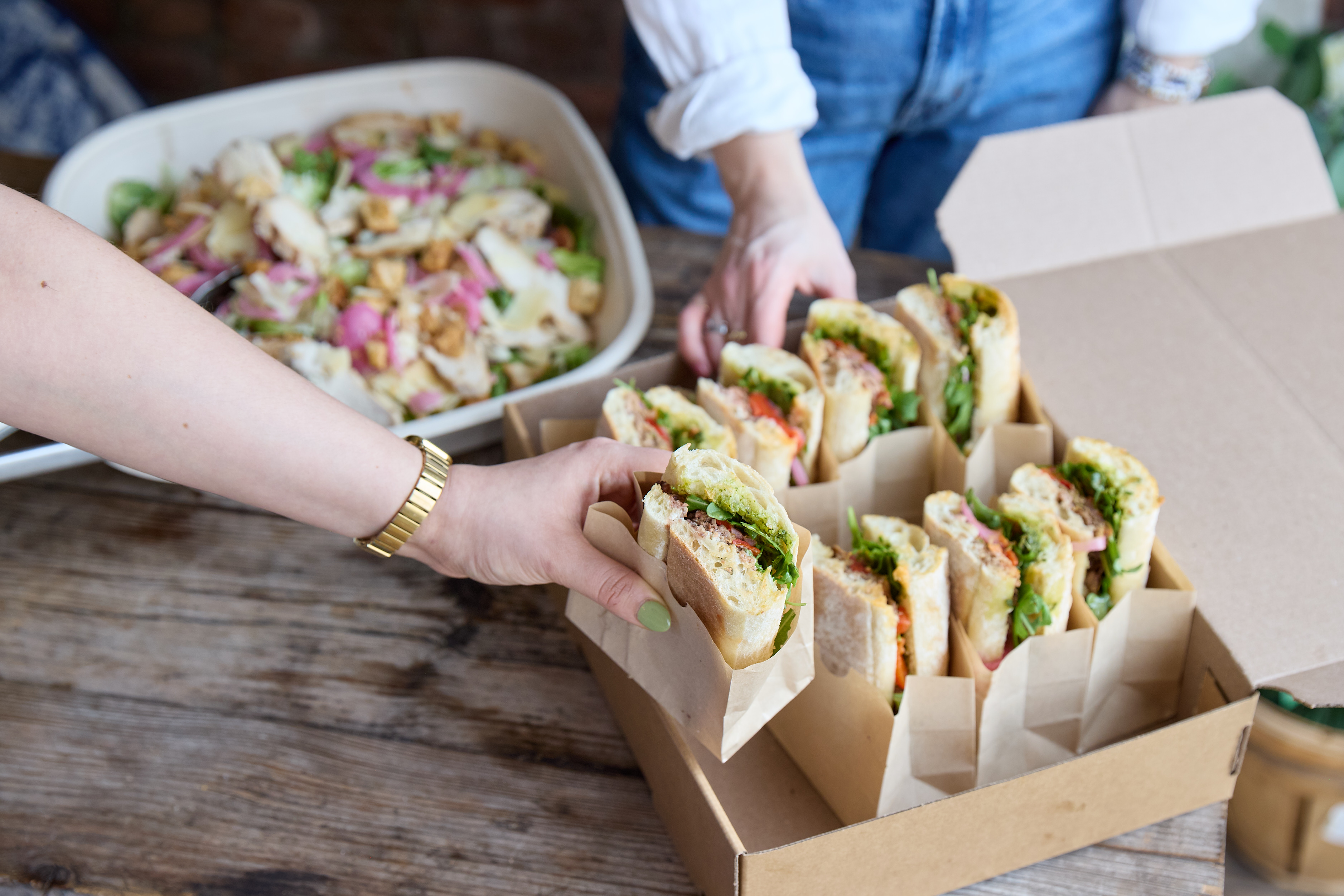 hands reaching for sandwiches packed in a brown catering box, with a large salad in the background on a rustic wooden table