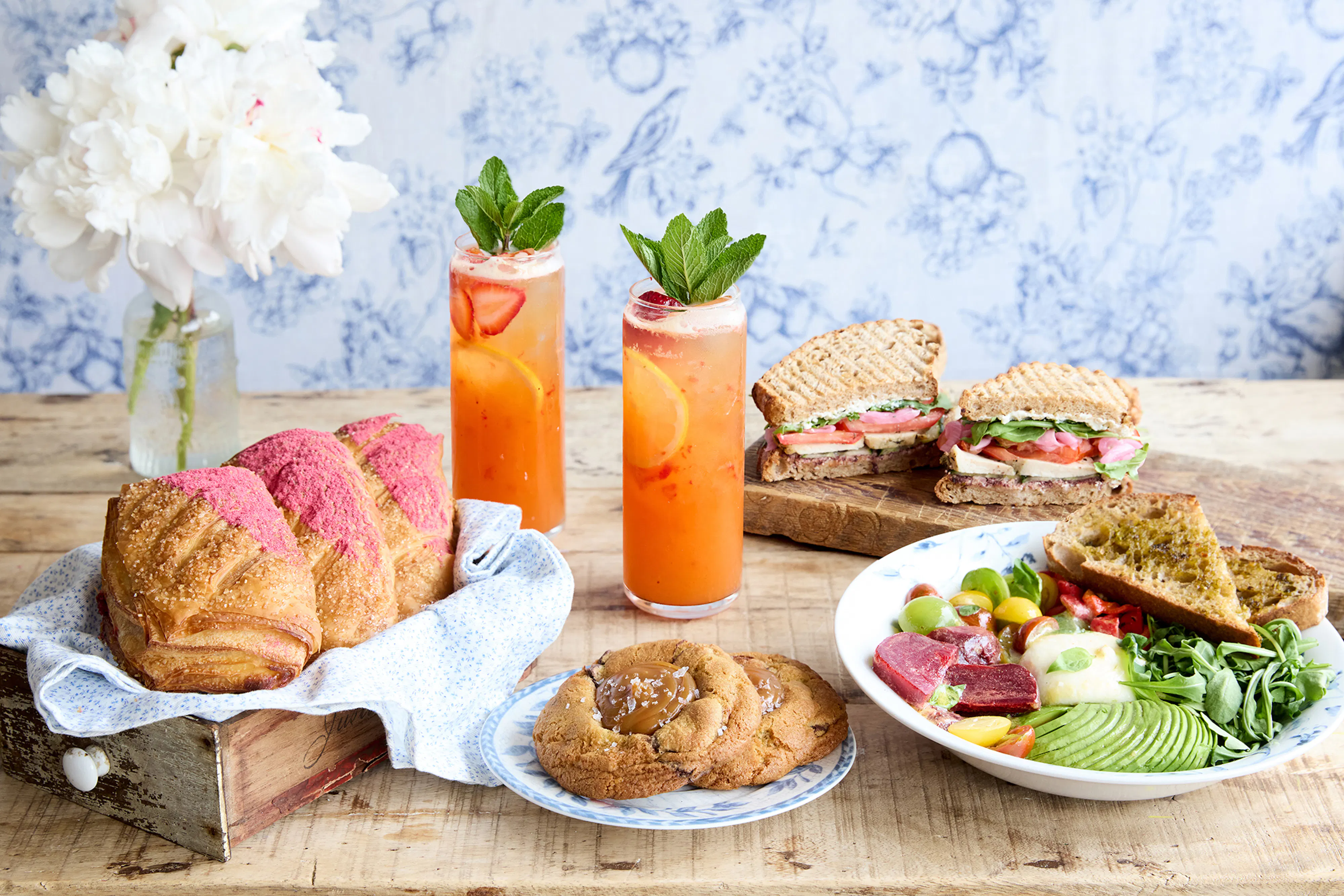 A summer table spread featuring Martha Stewart x Maman menu items, including vibrant beverages, a pink-glazed croissant, a fresh sandwich, seasonal salad, and cookies, styled against Maman's signature blue-and-white floral backdrop.