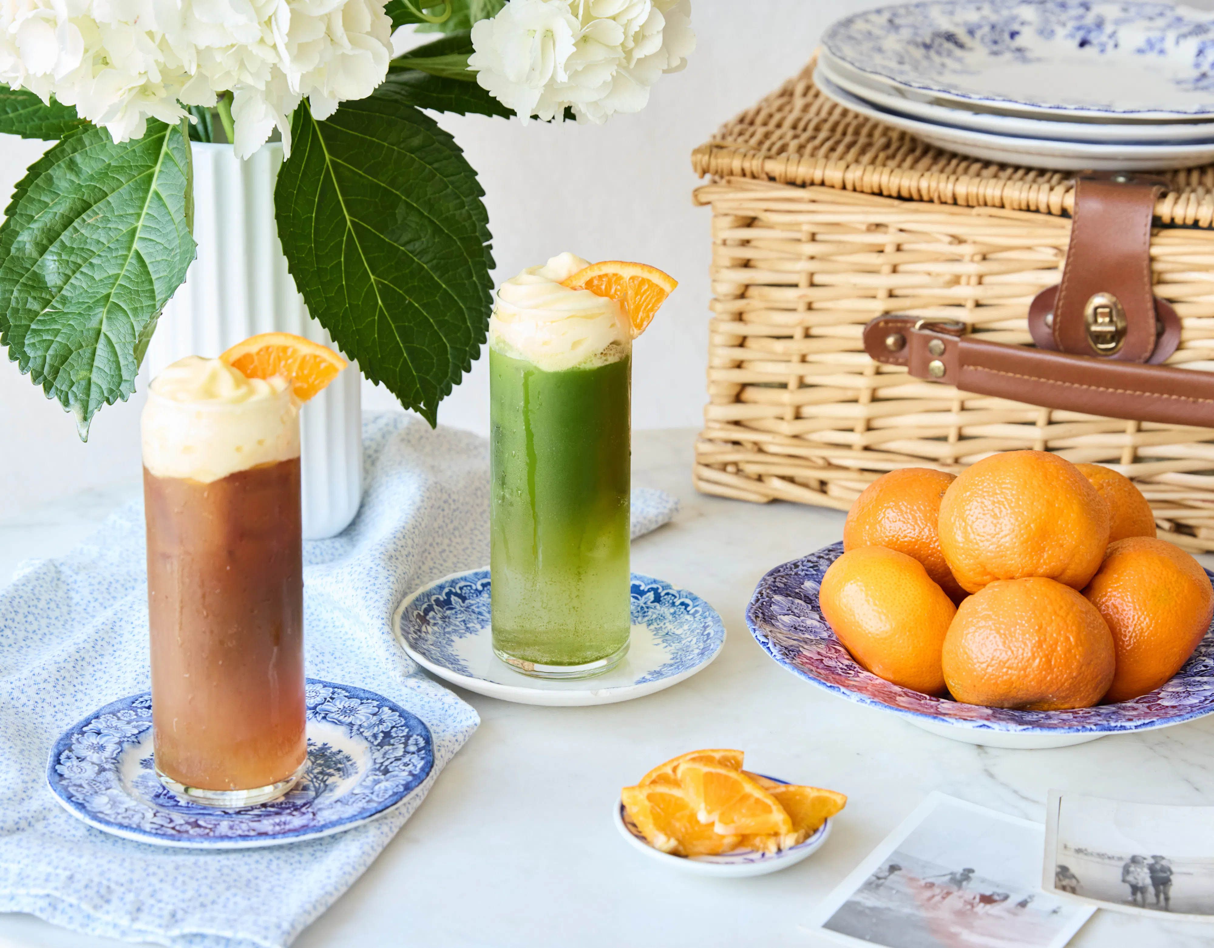 Two seasonal iced beverages are styled alongside fresh oranges, hydrangeas, and a picnic basket, capturing the bright, nostalgic spirit of summer.