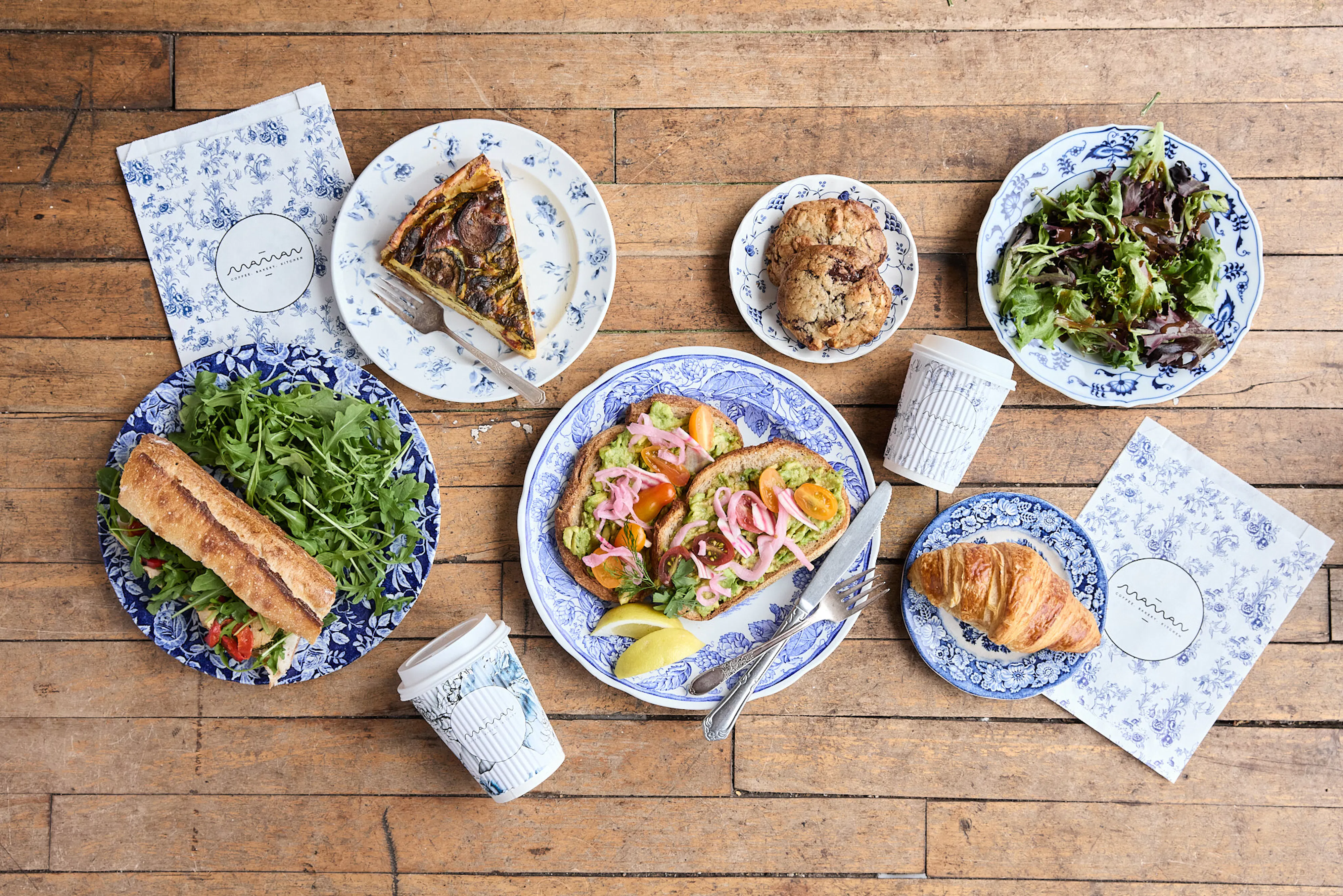 overhead shot of katie's chicken sandwich, quiche, avocado toast, croissant, cookies, and mixed greens with maman to-go cups and bags