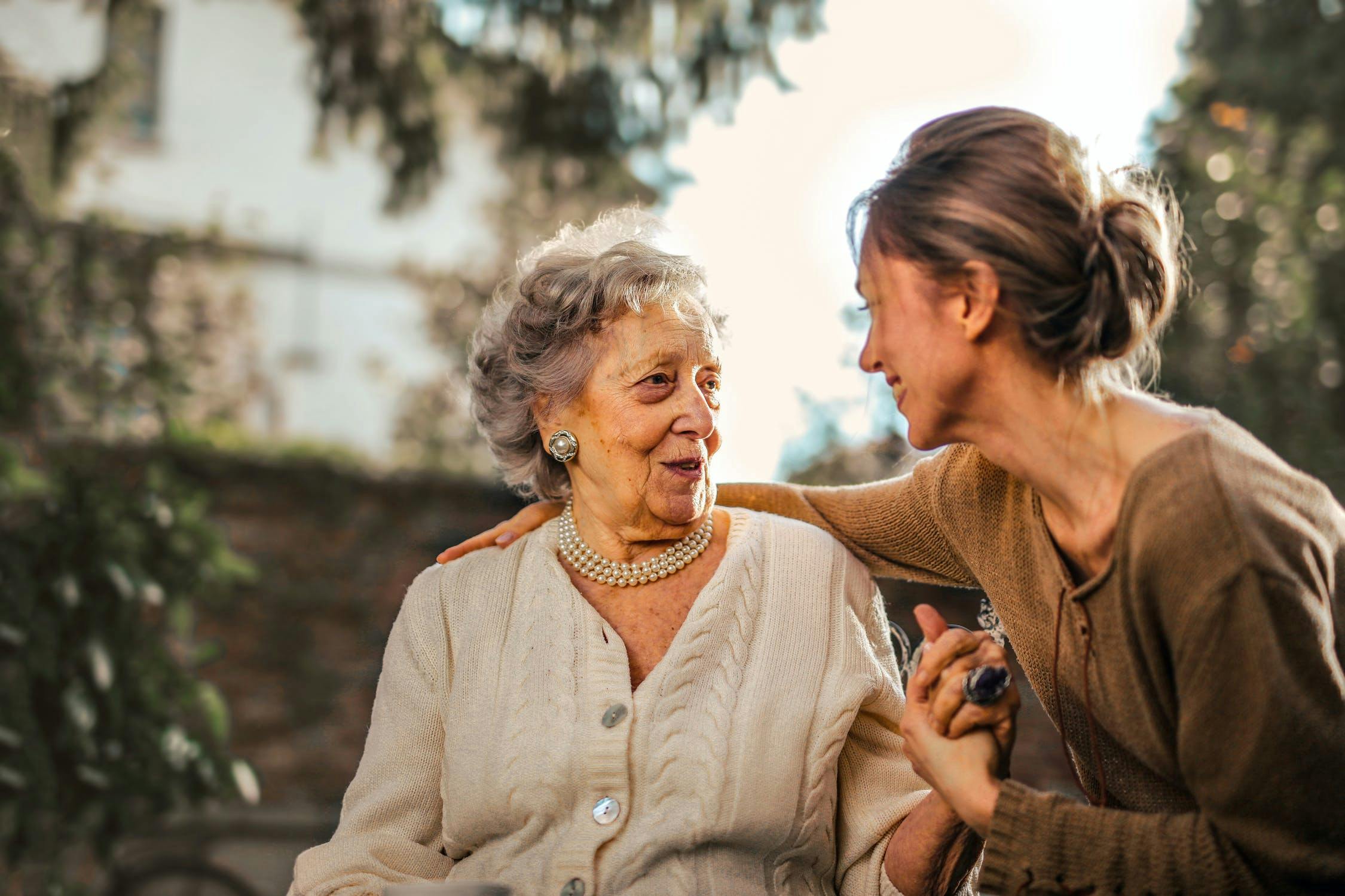 Elderly mother looking at adult daughter while smiling and holding hands.