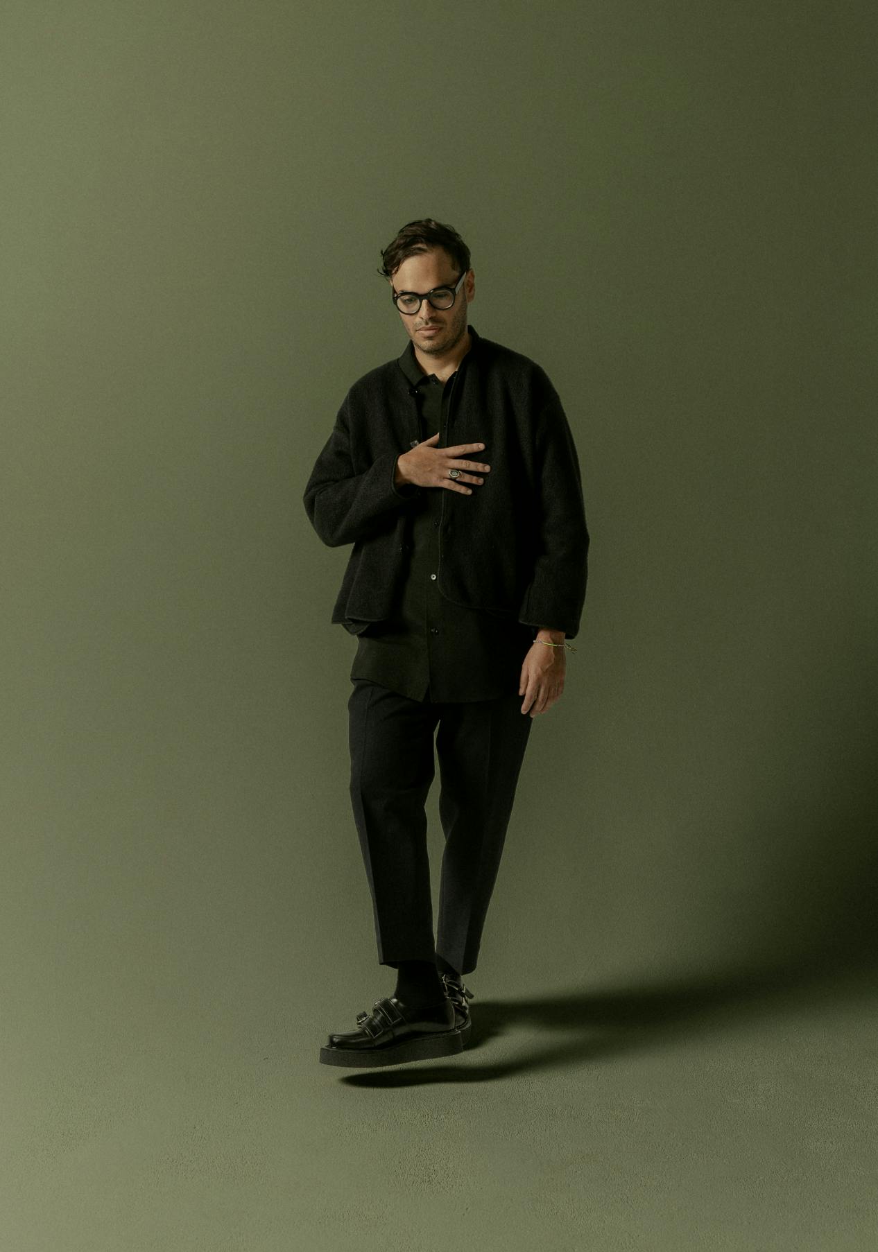 Bryan Senti holding his hand to his chest and stepping toward the camera in front of an olive-green background.