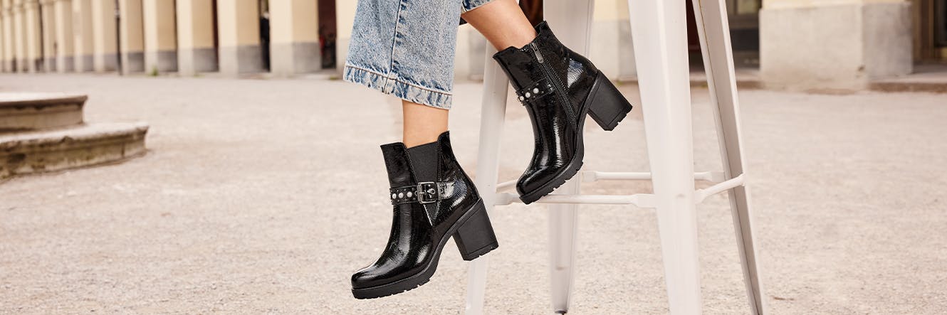 Frau mit hochgekrempelter Jeans sitzt auf einem weißen Stuhl und trägt glänzende schwarze Stiefeletten mit Blockabsatz und Schnallen.