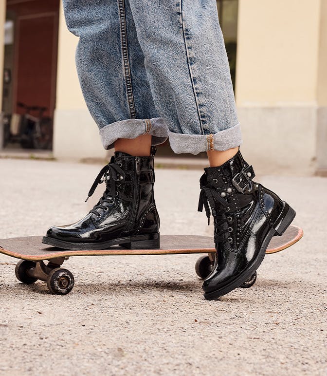 Donna in tuta di jeans e occhiali da sole accovacciata sul pavimento con uno skateboard, indossa stivali stringati neri lucidi con borchie e dettagli con fibbia.