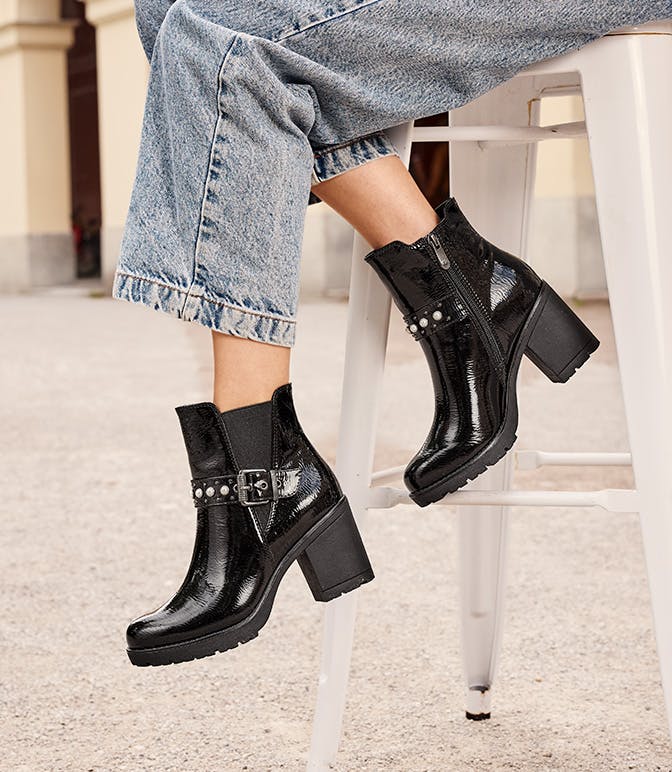 Stivaletti neri con tacco, borchie argentate e dettagli con fibbia, indossati con jeans larghi arrotolati su uno sgabello bianco.
