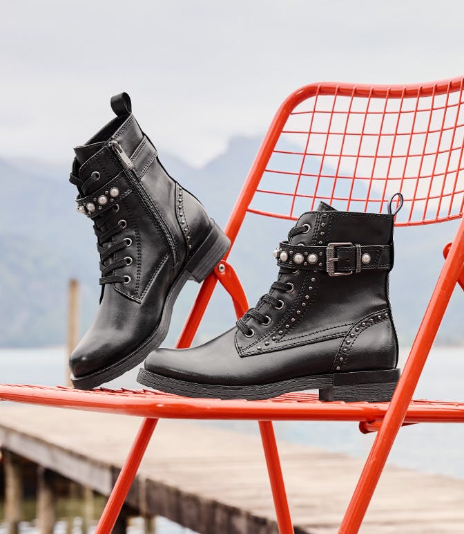 Coppia di stivali in pelle nera con lacci, borchie argentate e fibbia, disposti in modo decorativo su una sedia di metallo rosso davanti a uno sfondo di mare e montagne.