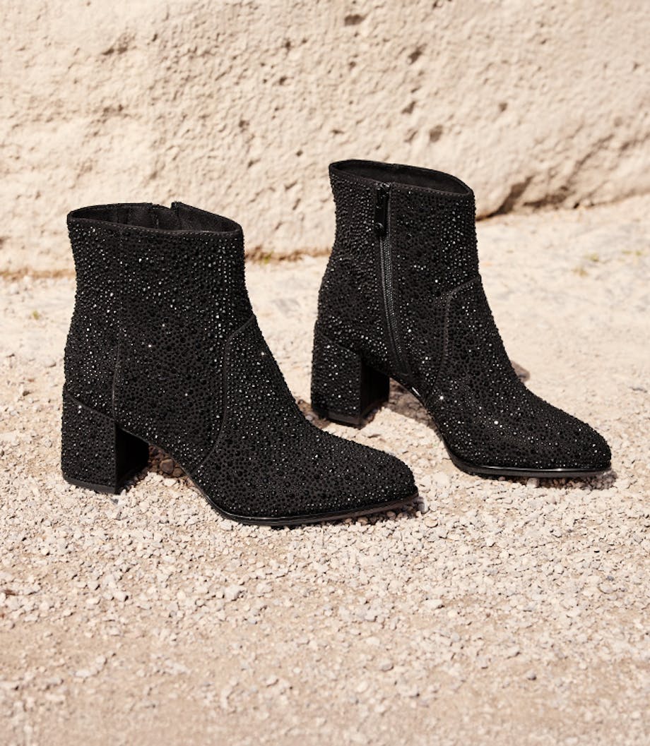 Dvě černé, třpytivé kotníkové boty s blokovým podpatkem stojí vedle sebe na světlém kamenném podkladu.