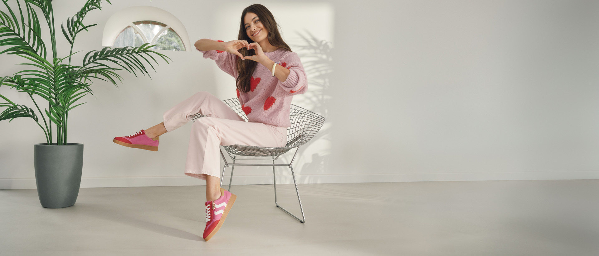 Una donna è seduta rilassata su una sedia moderna e forma un cuore con le mani. Indossa un maglione rosa con motivi a cuore, pantaloni chiari e sneakers rosa di MARCO TOZZI in una stanza luminosa e accogliente.