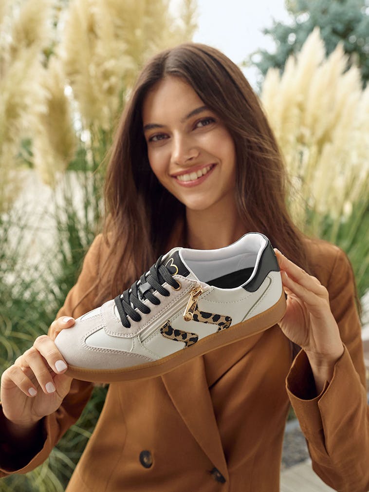 Lächelnde Frau im braunen Blazer präsentiert einen weißen Sneaker mit schwarzer Schnürung, leo-gemusterten Details an der Seite und goldfarbenem Reißverschluss vor hellem Naturhintergrund