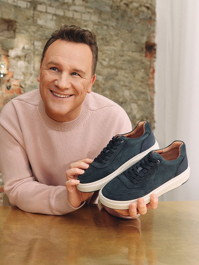 Guido in rosafarbenem Pullover präsentiert dunkelblaue Sneaker mit weißer Sohle und Schnürung auf einem Holztisch vor Ziegelwand