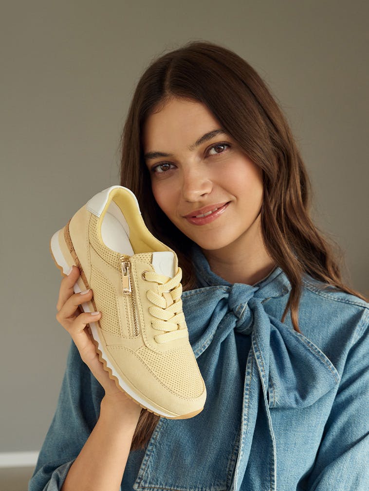 Frau mit braunen Haaren hält gelben Sneaker mit Reißverschluss und Schnürung neben ihr Gesicht, trägt blaue Bluse mit Schleife