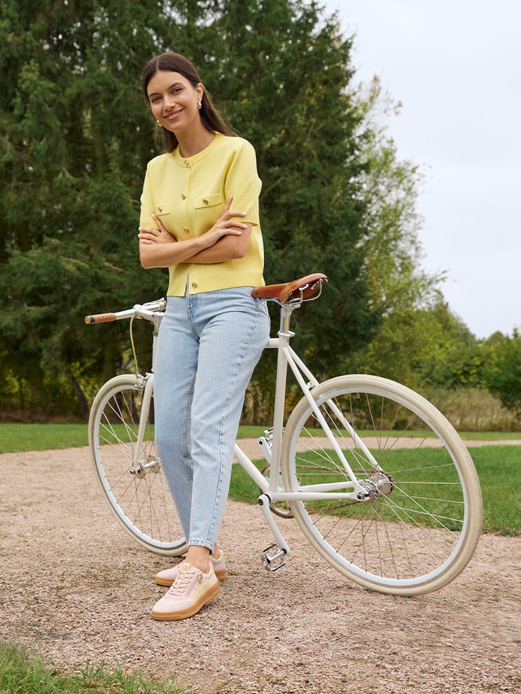 Frau lehnt an einem Fahrrad auf einem Parkweg und trägt Sneaker in Rosa und Beige zu Jeans und gelber Strickjacke. 