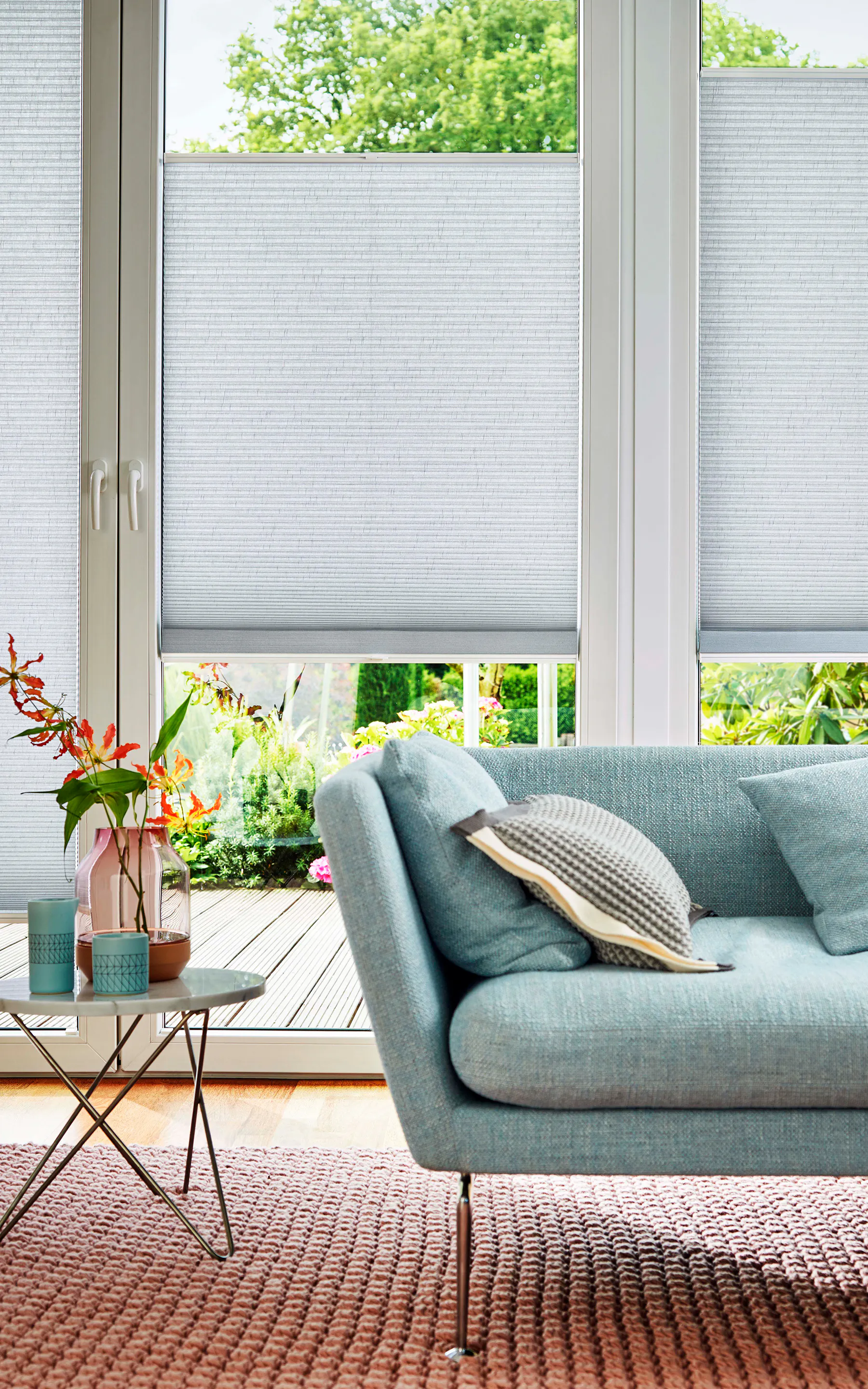 Plisségardiner er delvis trukket ned over store glassdører, skjermer innsyn. Foran står en lyseblå sofa med puter, et lite bord med vaser, og en grønn hage utenfor.
