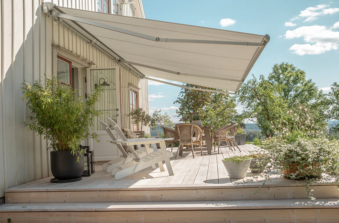 En foldbar hvit markise strekker seg ut og gir skygge over en treveranda med stoler og et spisebord, omgitt av potteplanter og trær under en lys, solrik himmel.