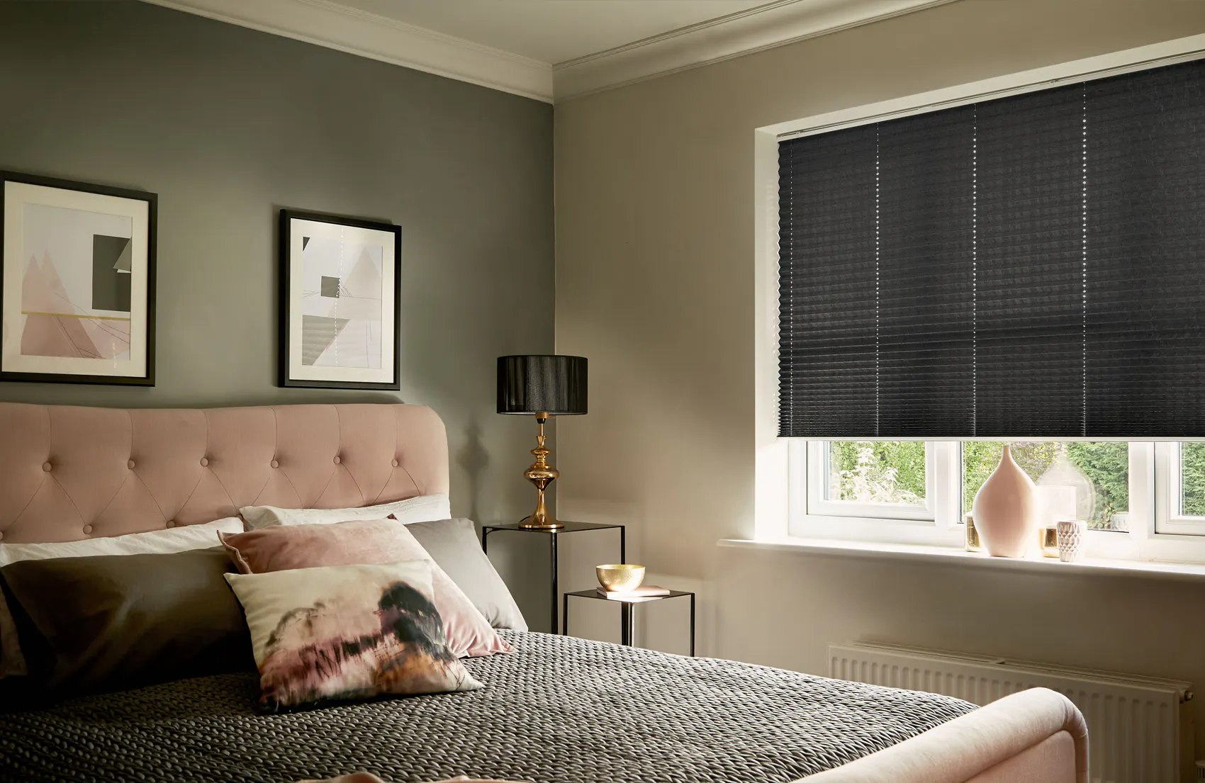 Mørkt plisségardin er trukket ned over vinduet og demper dagslys i et dempet soverom med rosa sengegavl, nattbord med lampe og to innrammede abstrakte bilder.