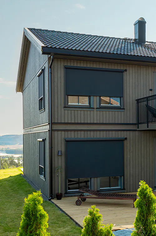 Mørke utvendige rullegardiner dekker to vinduer i en moderne trefasadebolig; ett fullstendig nede og ett delvis nede, ved balkong og landskapsutsikt mot innsjø.