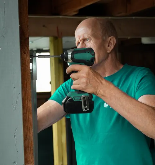 En mann monterer en persienne med en batteridrevet drill i en tre vindusramme innendørs, konsentrert arbeid ved et vindu i et uferdig eller oppussingsrom.