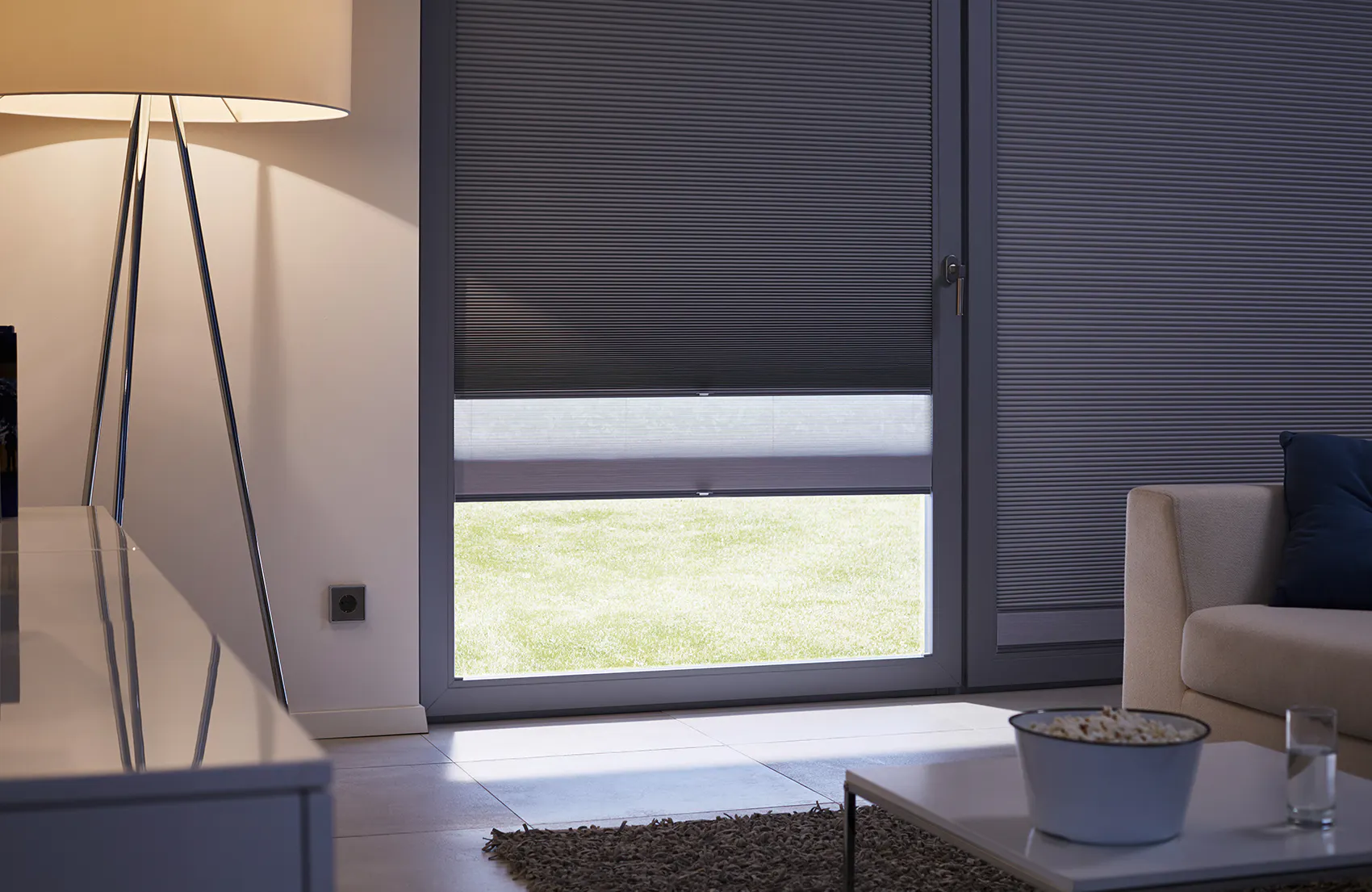 Plisségardin delvis senket over vindu, blokkerer lys i øvre del og lar et lysere panel vise gress utenfor. Stue med sofa, gulvlampe, salongbord og teppe.