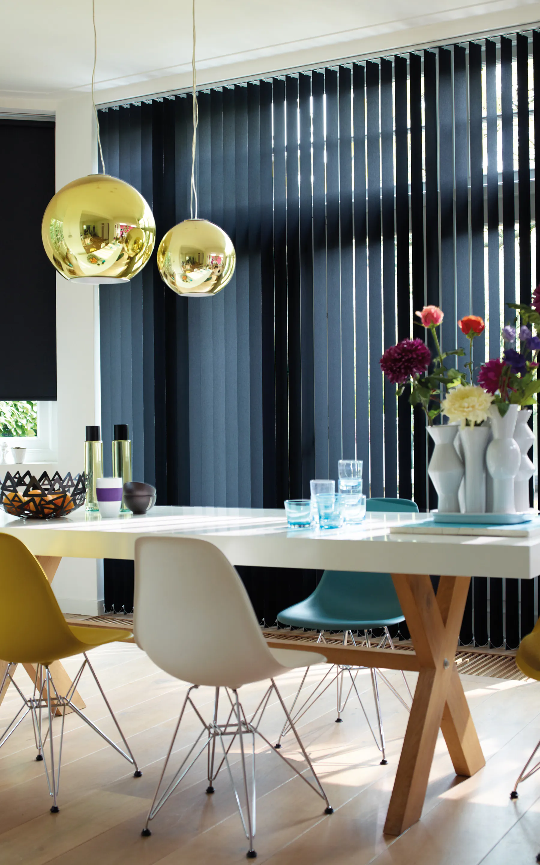 Mørkeblå vertikal lamellgardin filtrerer dagslys og danner stripete lys over et lyst spisebord. Moderne spisestue med fargerike stoler, gullpendellamper, glass og vase med friske blomster.