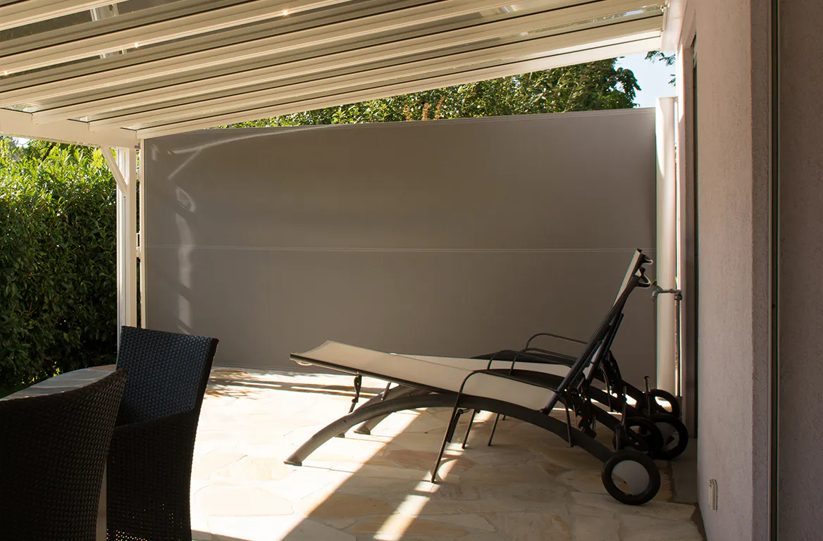 Grått utendørs rullgardin/sidevegg er trukket ut og gir skjerming og skygge på en steinlagt terrasse under en pergola, med to solsenger og hagemøbler i bakgrunnen.