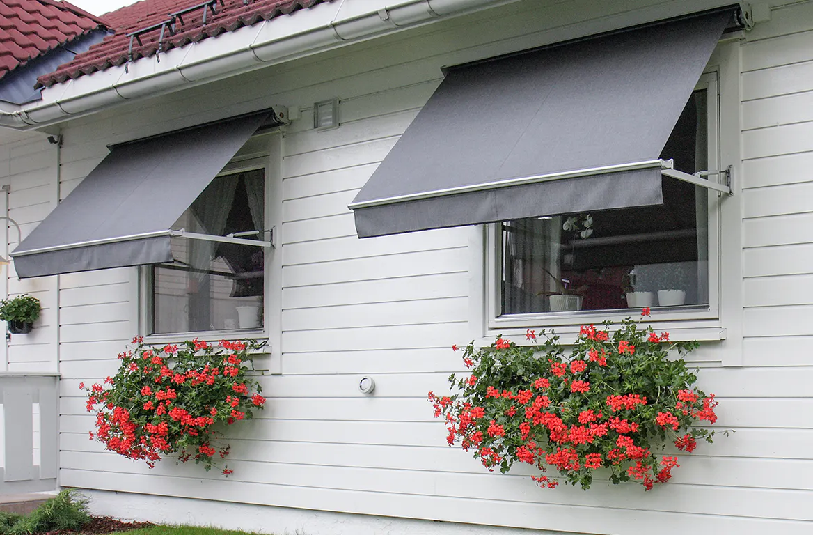 To grå, uttrukne markiser skjermer to vinduer i en hvit liggende trepanelfasade; under hvert vindu henger frodige røde pelargonier i blomsterkasser. Rødt tegltak og grønn plen rundt.