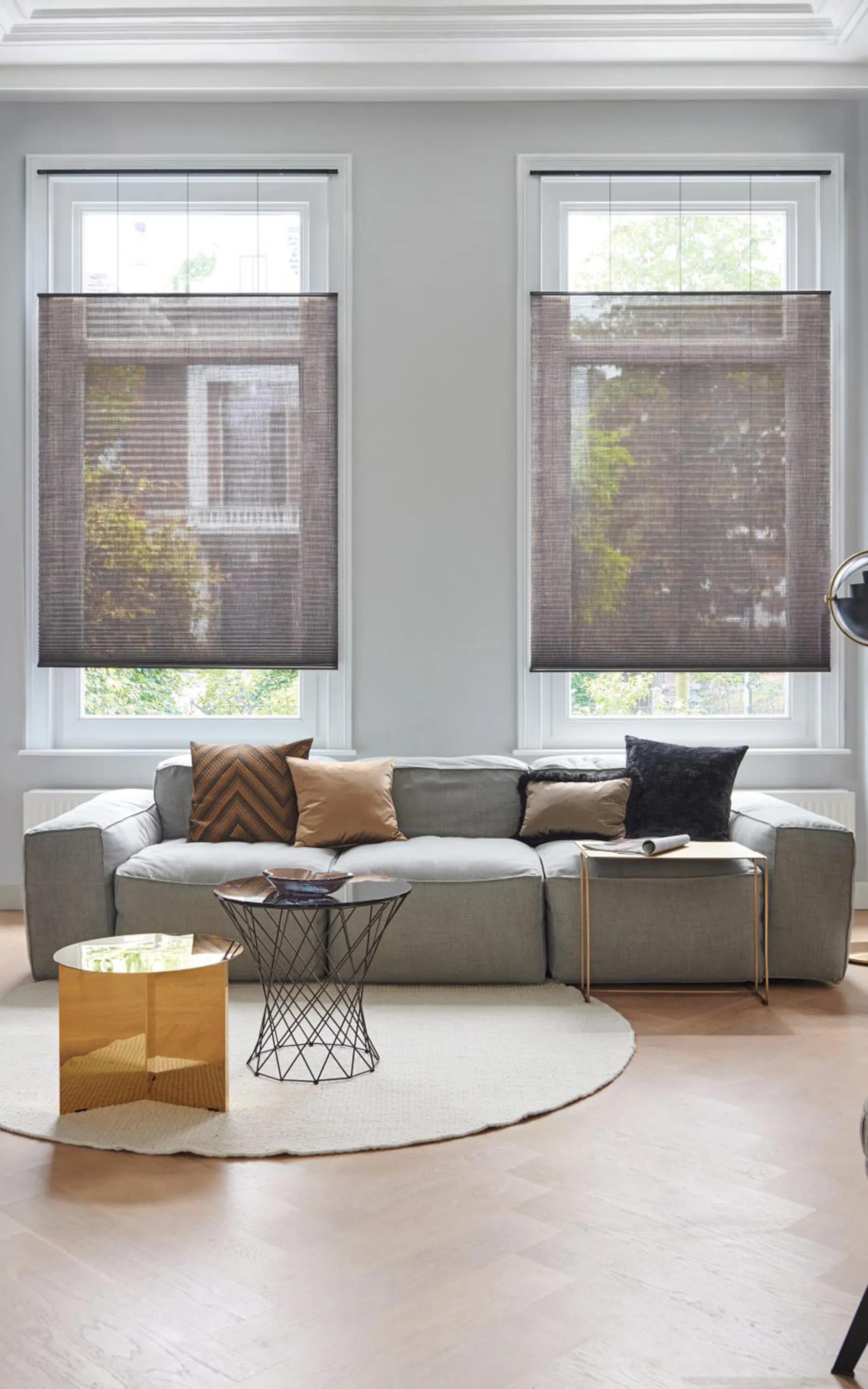 To grå plisségardiner filtrerer dagslys i et lyst, moderne oppholdsrom; foran dem står en lav grå sofa med flerfargede puter, rundt teppe og to moderne småbord.