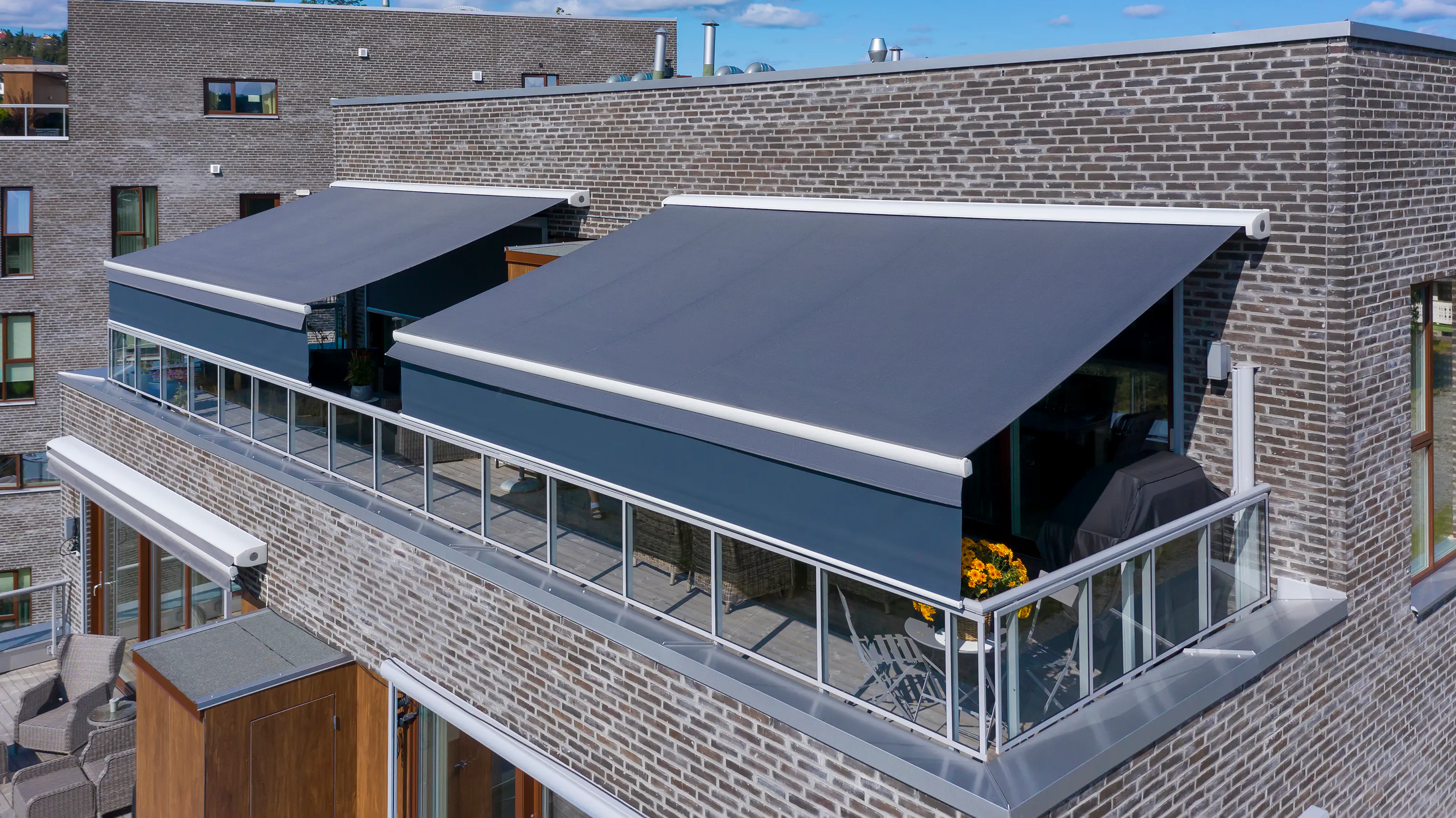 Mørkegrå terrassemarkiser er trukket ut og gir skygge over balkonger på en moderne mursteinsbygning med glassrekkverk, hagemøbler og blomsterkasse.
