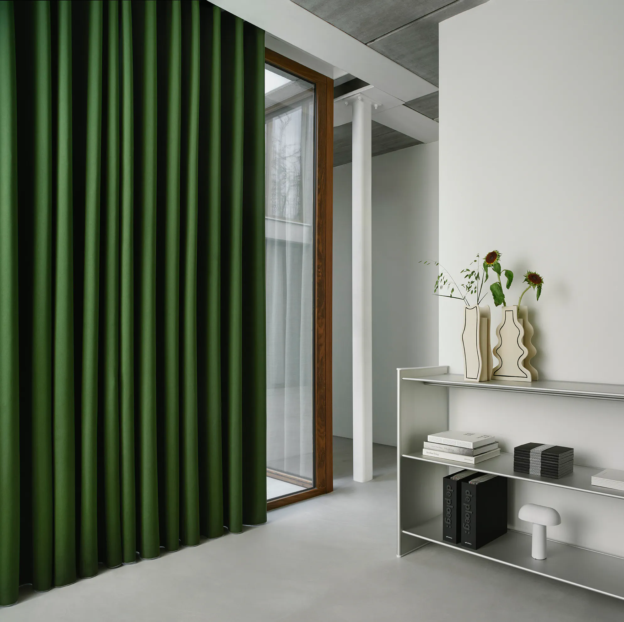 Mørkegrønne, tunge gardiner henger delvis foran et høyt glassdørvindu i et lyst, minimalistisk interiør med hvite vegger, søyle og en metallhylle med vaser og bøker.