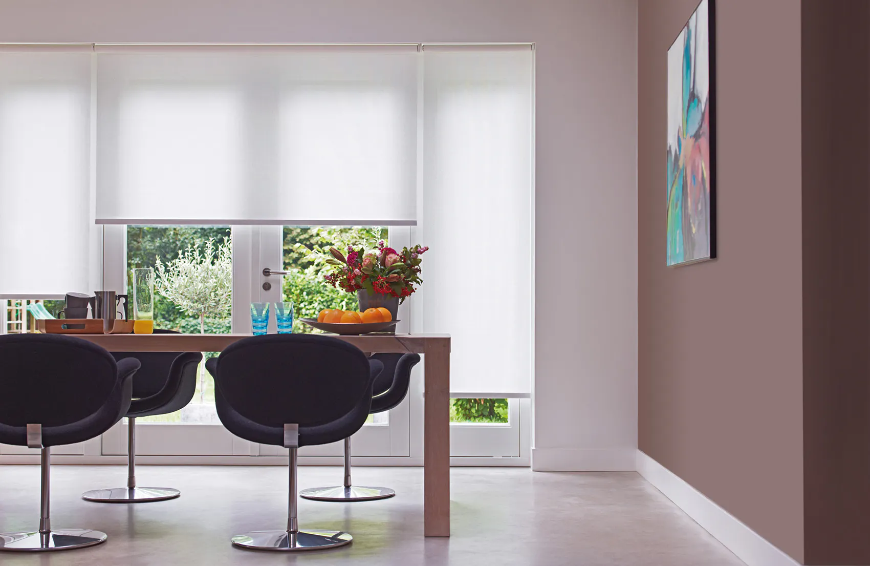Hvite rullegardiner dekker store vinduer og filtrerer dagslys; et trebord med fire moderne svarte spisestoler, frukt og vase står foran vinduet i en lys, minimalistisk stue med veggkunst.