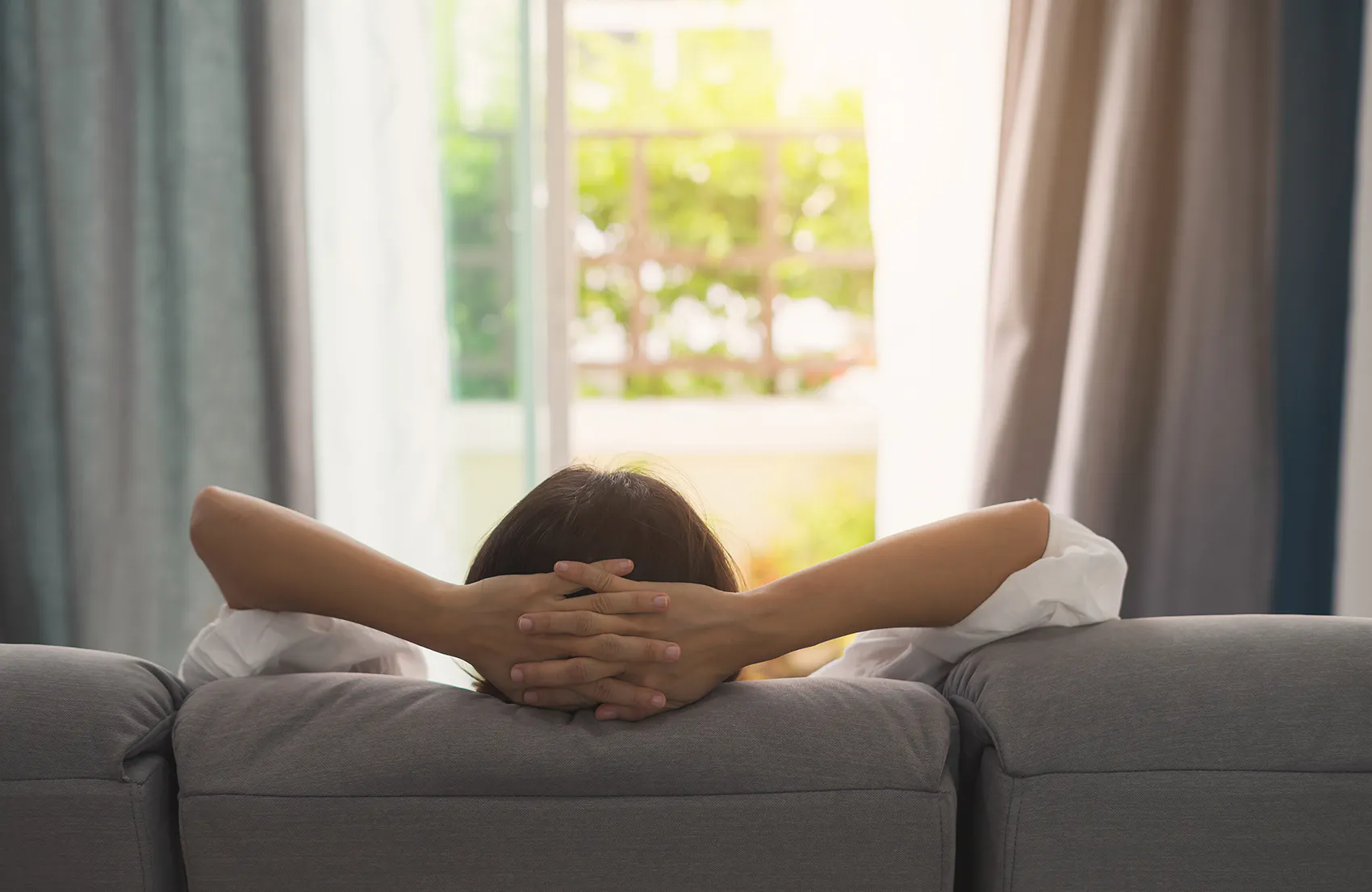Grå gardiner rammer et lyst vindu og slipper inn dempet dagslys; en person sitter i sofaen med hendene bak hodet og slapper av i en rolig stue.