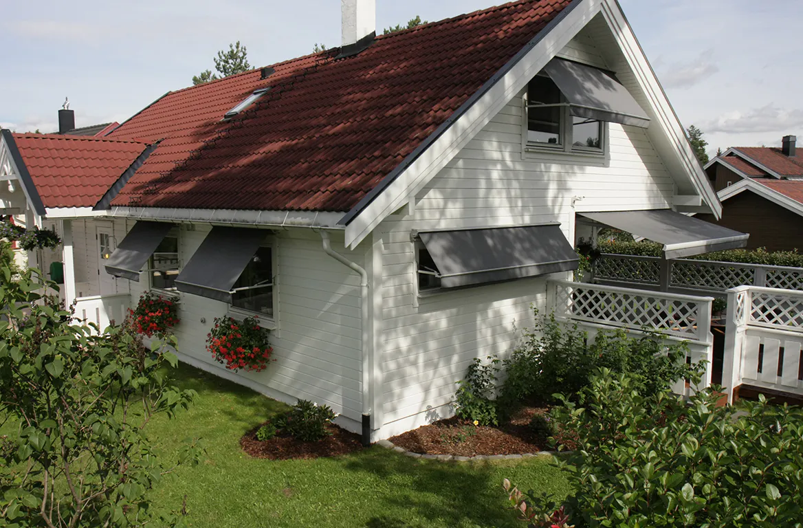 Utvendige grå markiser dekker og skygger flere vinduer og en liten veranda på et hvitt, skråtaket hus med rødt tak. Omgitt av grønt gress, blomsterkasser og hageplanter på en solrik dag.