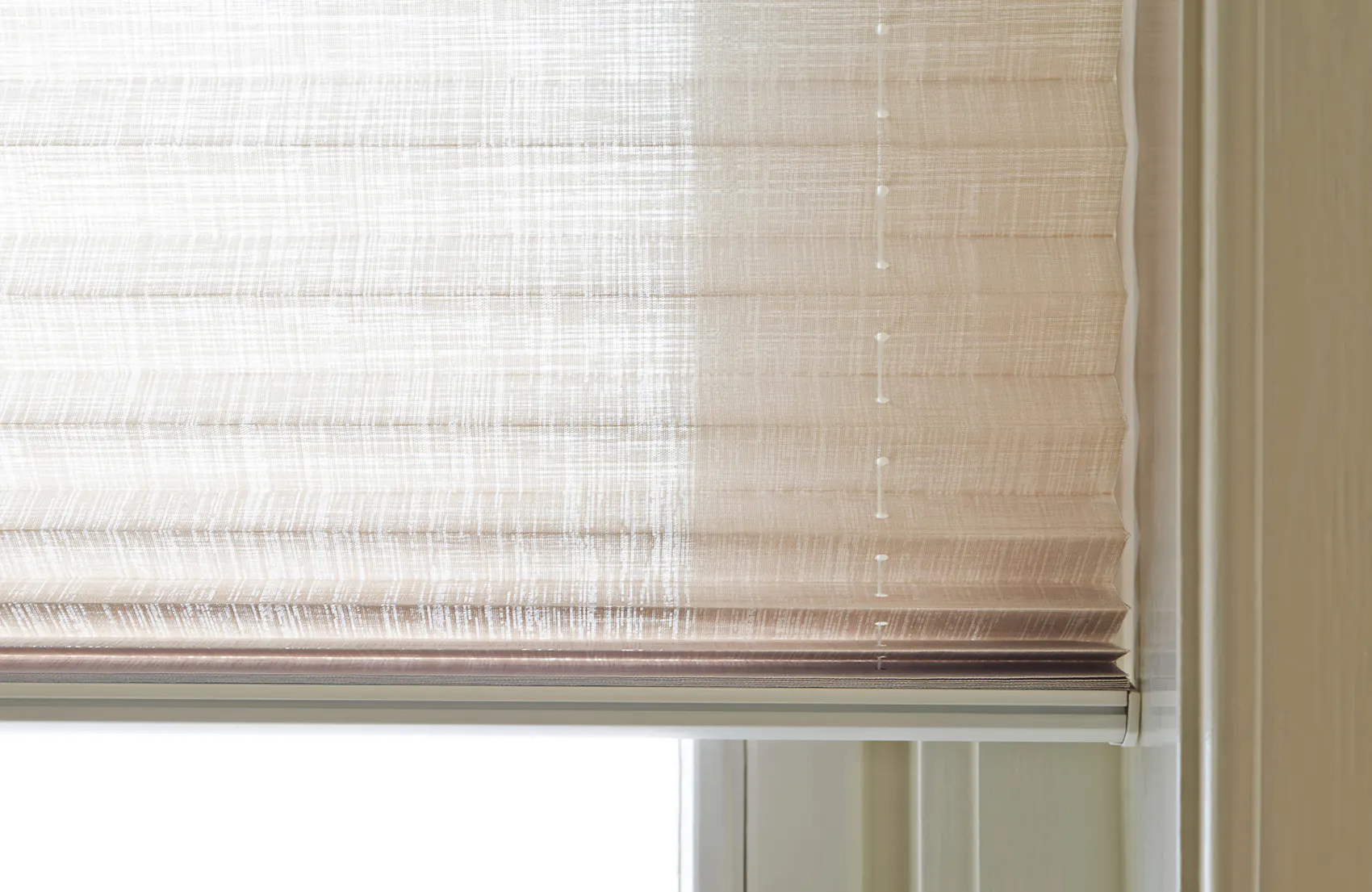 En kremfarget, halvsenket foldgardin (roman blind) dekker et vindu, filtrerer mykt dagslys inn og hviler innenfor en hvitmalt vinduskarm i et lyst interiør.