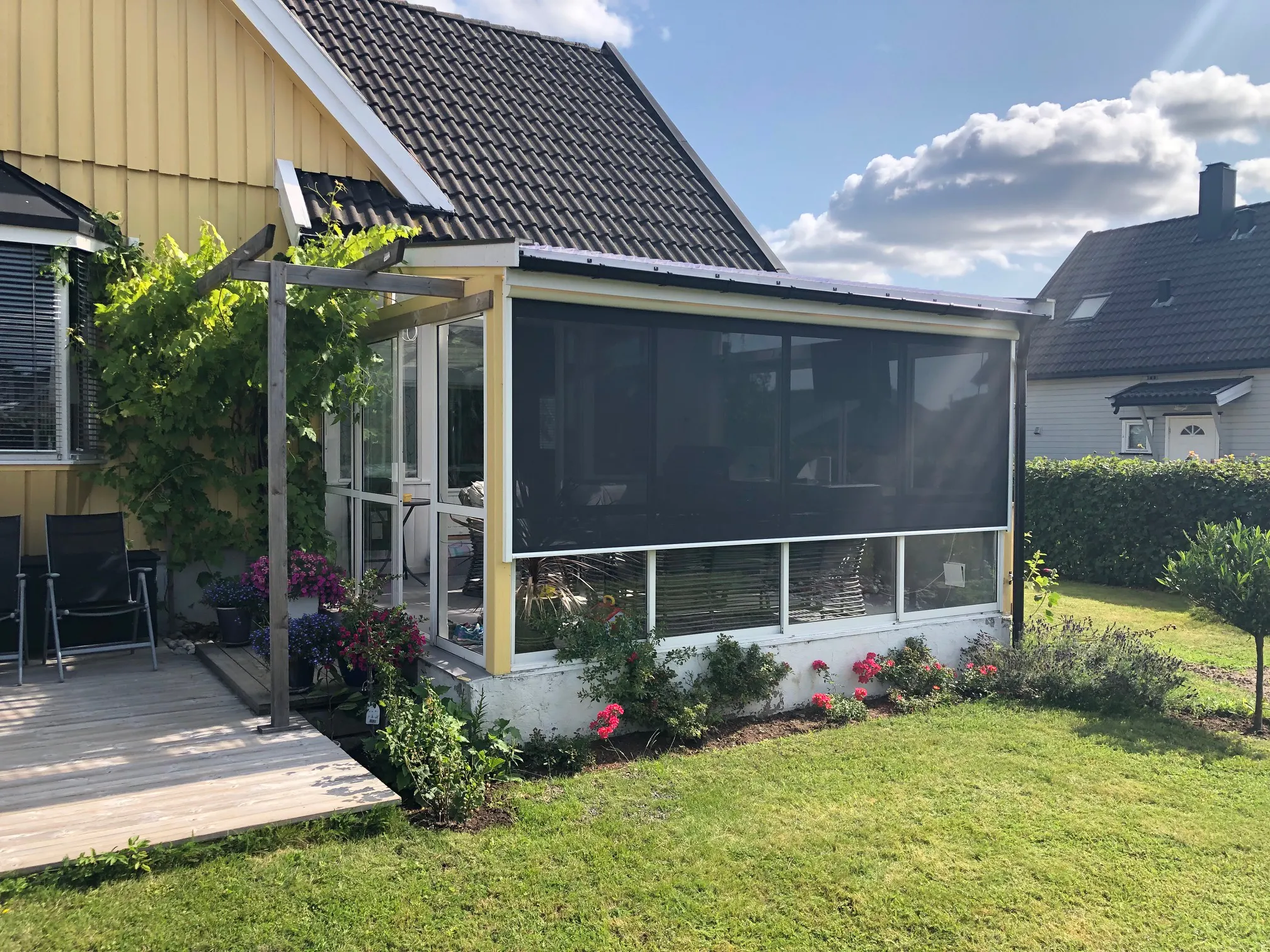 Mørk utvendig rullegardin dekker en innglasset veranda, skaper solskjerming og privatliv; montert på et gult hus med treterrasse, blomsterbed og grønn plen under en skyfri, solrik himmel.