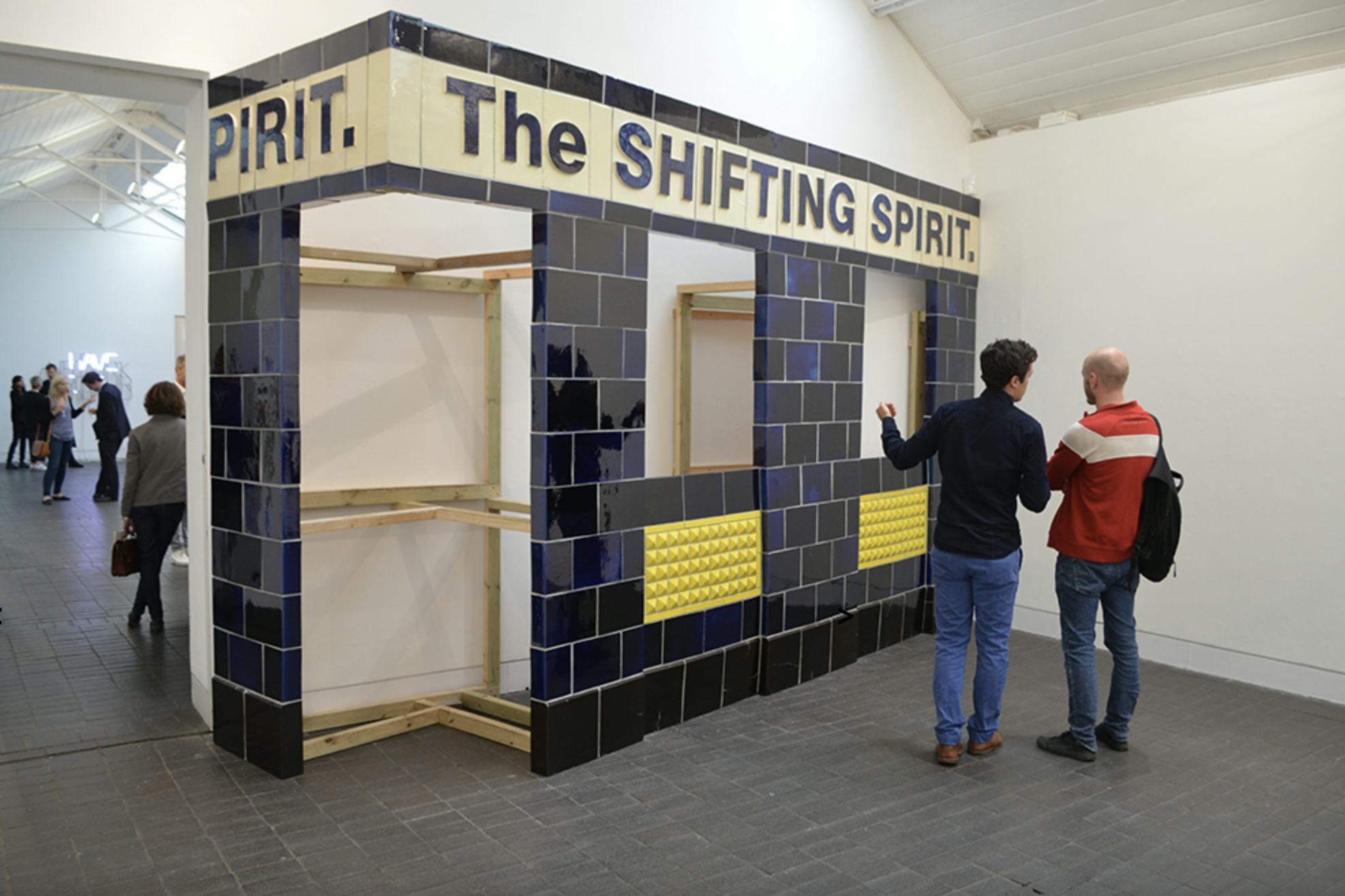 Two people stand in front of an artwork of a tiled facade of a pub. The text ‘The SHIFTING SPIRIT’ is visible on the artwork. More people are visible through a doorway into the next room of the art gallery