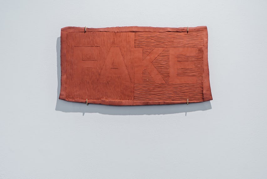 A ceramic artwork hanging on a gallery wall. The letters F A K E are impressed into the clay