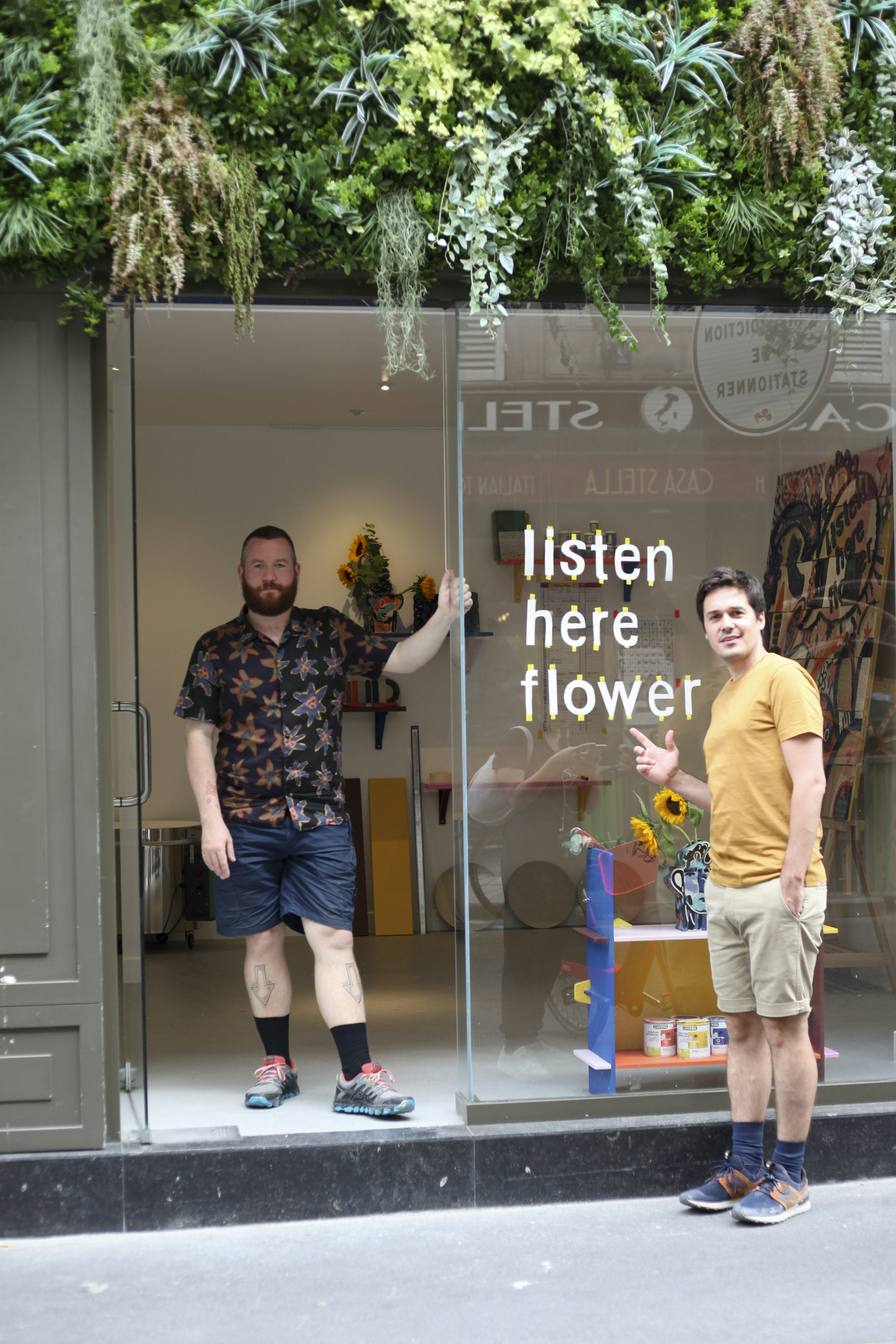 John Booth and Matthew Raw stand in the entrance of a gallery with the words ‘listen here flower’ on the window