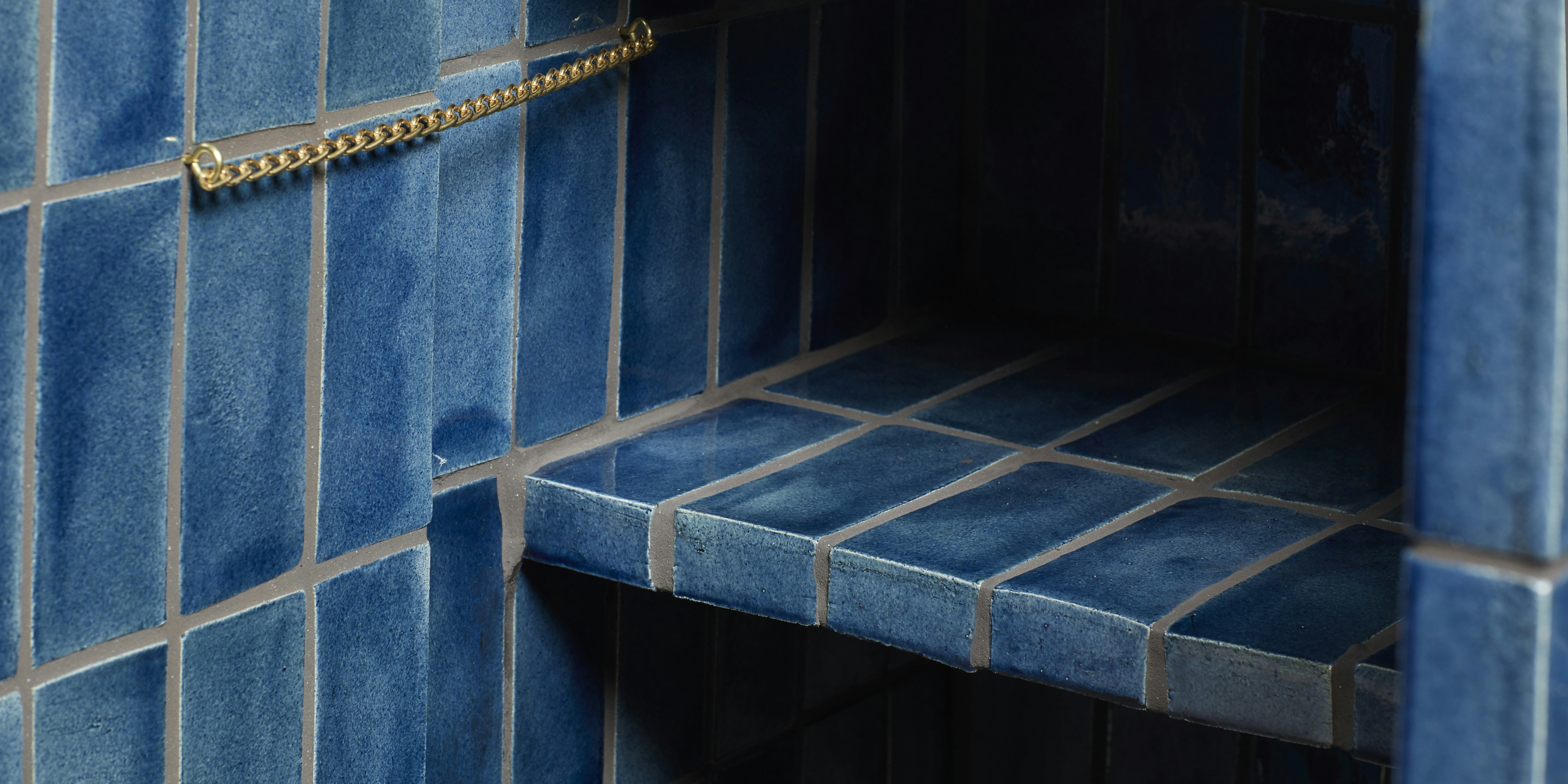 A detail of the inside of a sideboard clad in handmade blue tiles. A gold chain hangs between the inside of the sideboard and the door