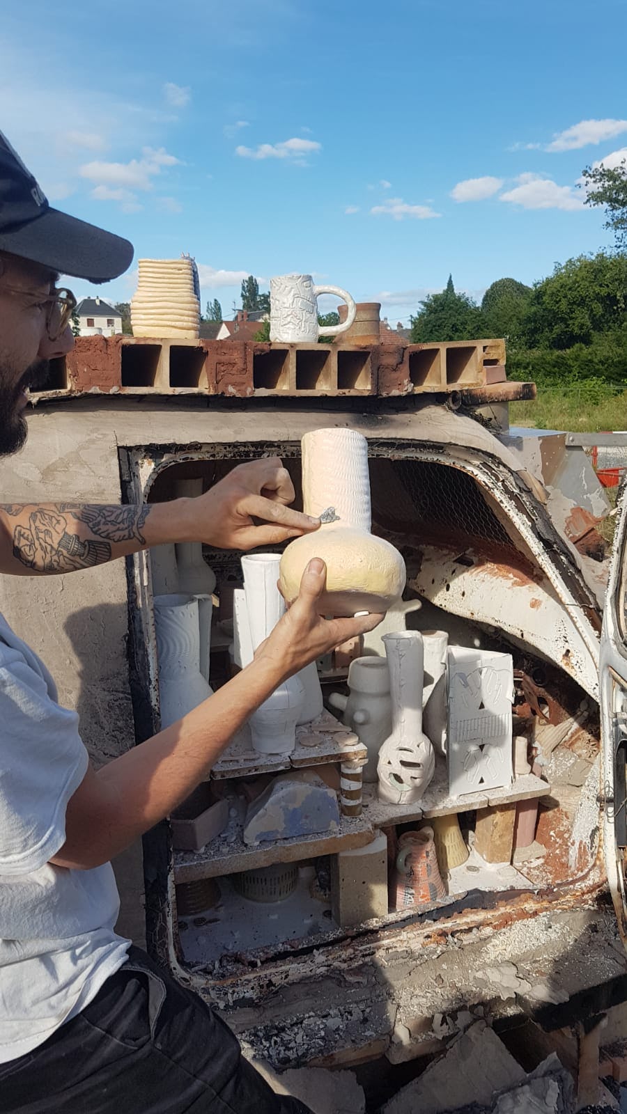 A man holds a ceramic vase in front of an old car filled with more ceramics