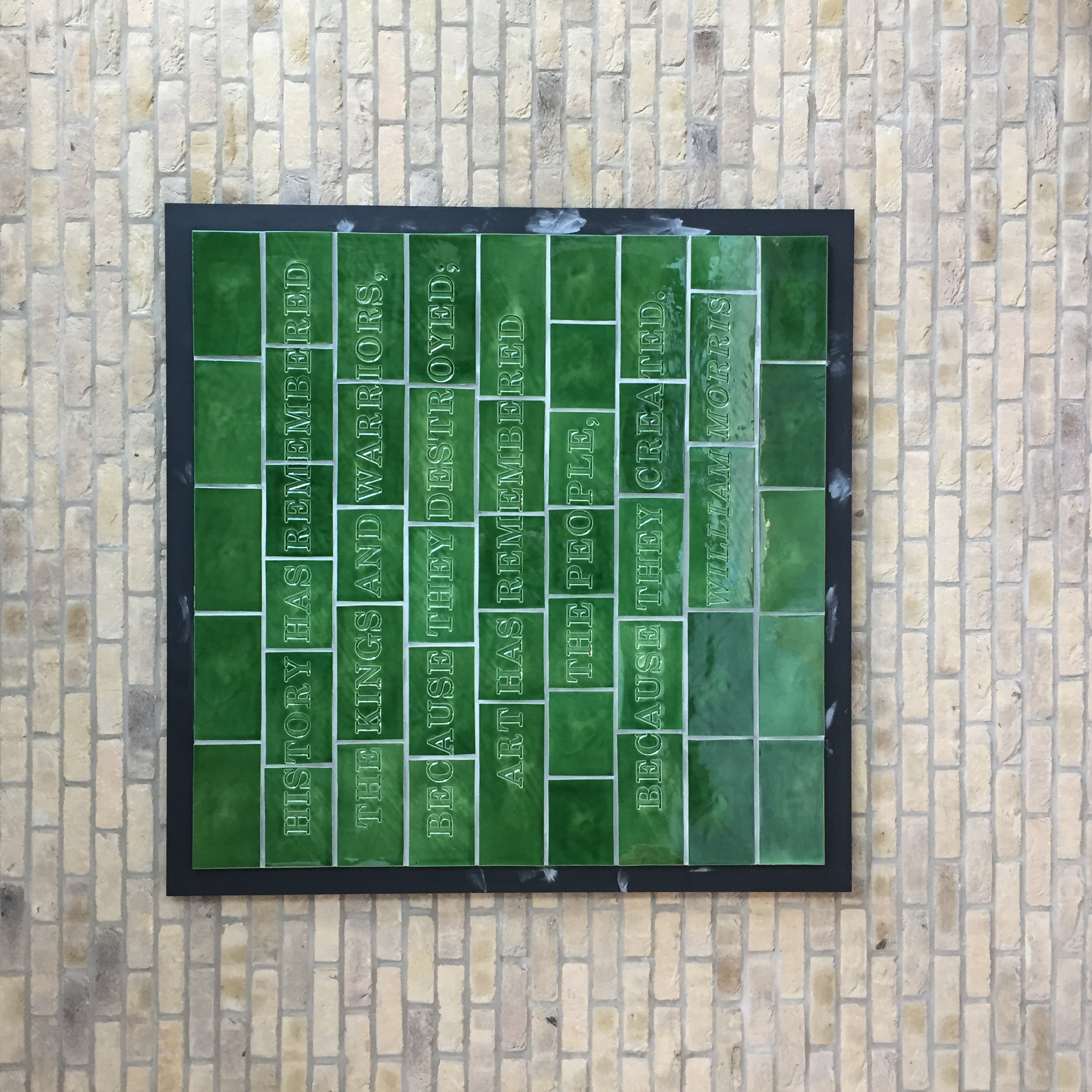 A green tiled artwork hangs on a brick wall. The text ‘History has remembered the kings and warriors, because they destroyed; art has remembered the people, because they created. William Morris’ is debossed into the tiles