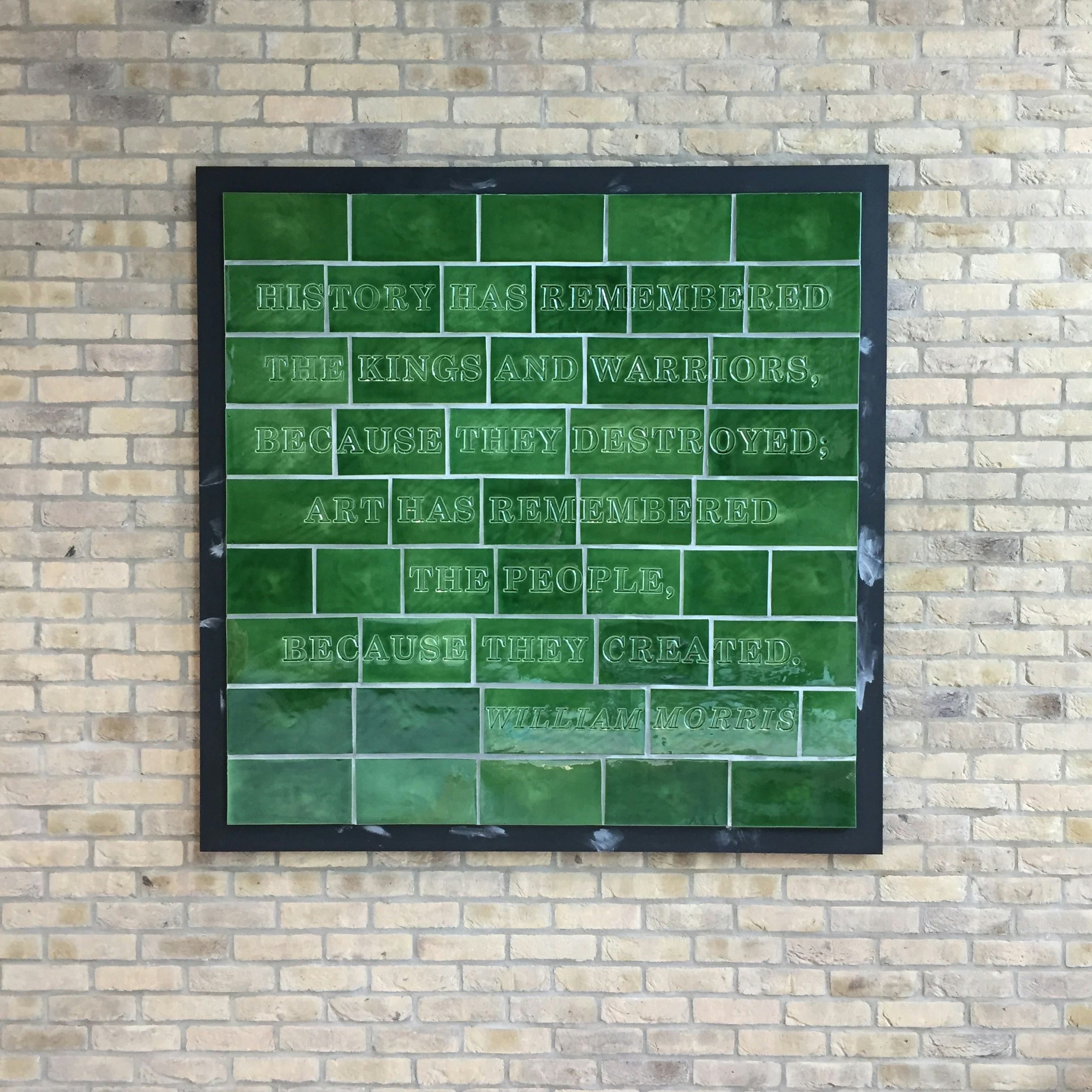 A green tiled artwork hangs on a brick wall. The text ‘History has remembered the kings and warriors, because they destroyed; art has remembered the people, because they created. William Morris’ is debossed into the tiles