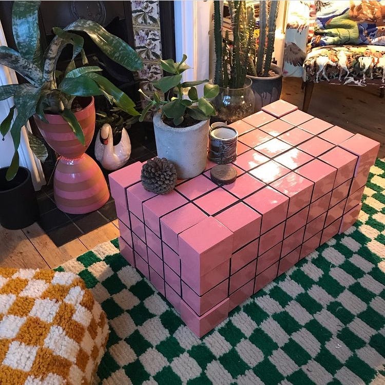 A table covered in pink tiles in a home with patterned rugs and plants in front of a fire place
