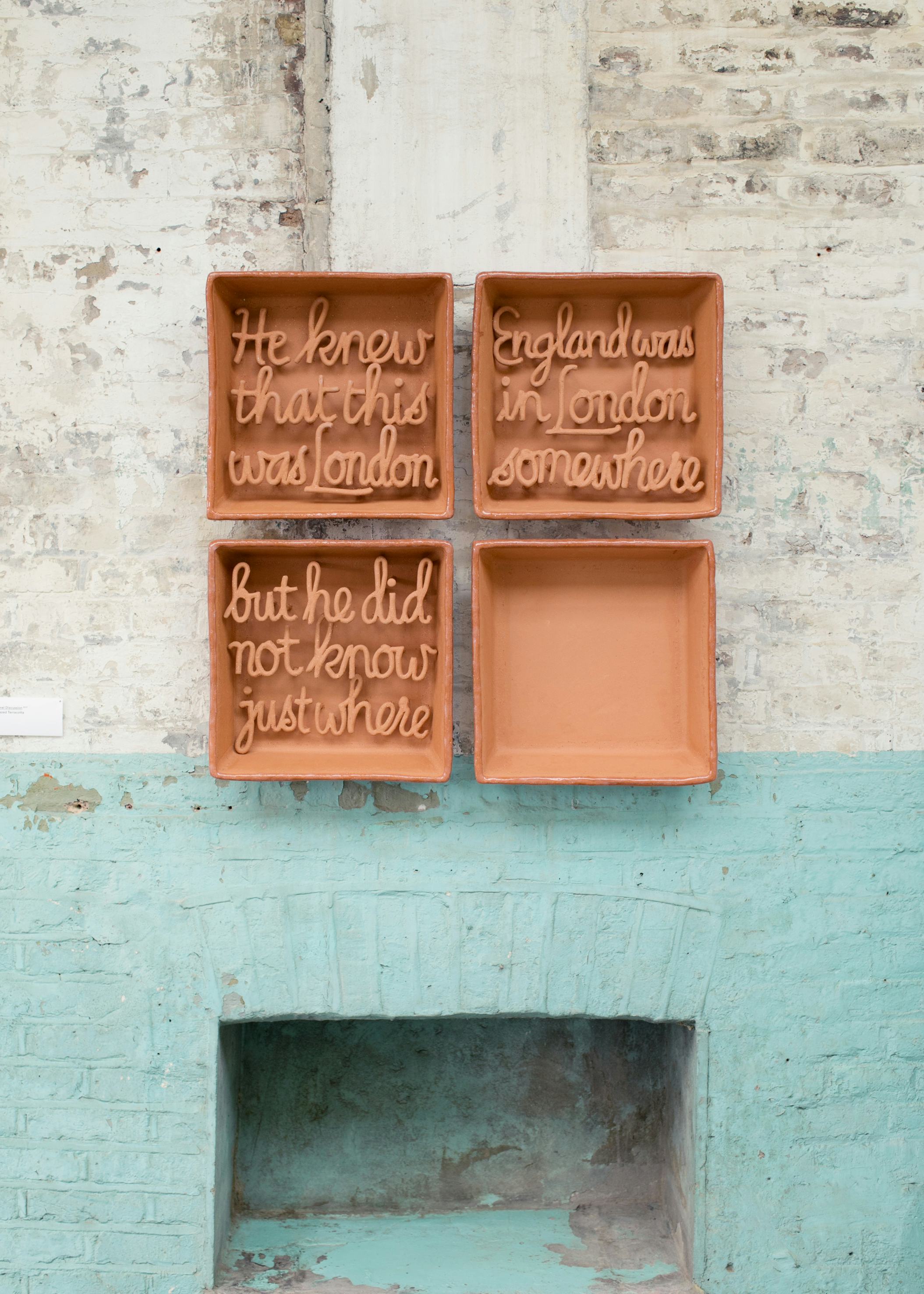 A ceramic artwork in four pieces hanging on a painted brick wall above an old fireplace. The artwork includes the text ‘He knew that was London. England was in London somewhere. But he did not know just where’