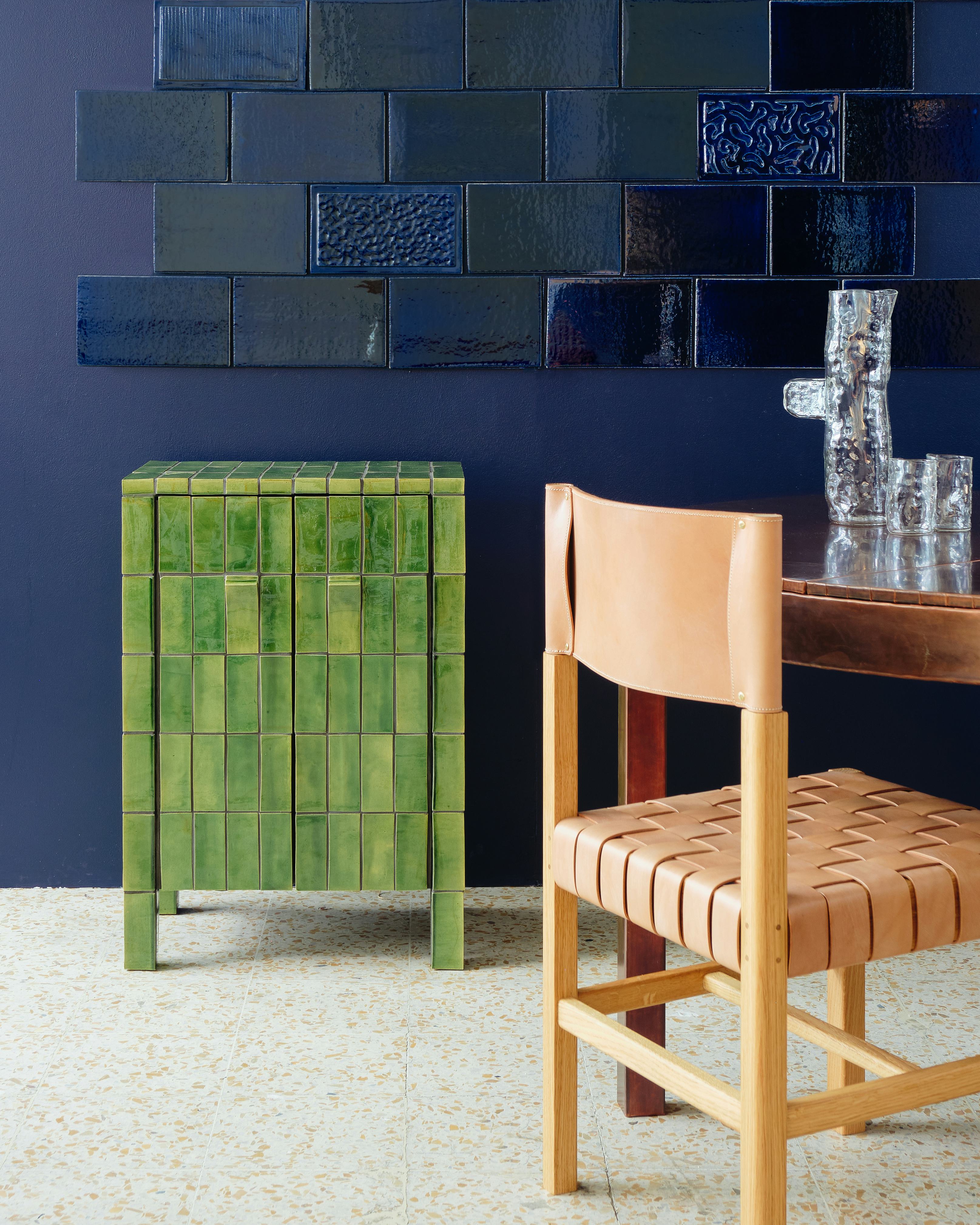 A green tiled cupboard in front of a blue tiled wall and next to a table and chair