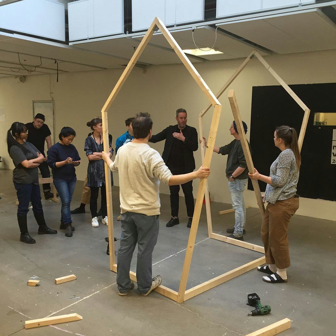 A group of people in a room. Three of them are holding a wooden structure in a the shape of a simple building