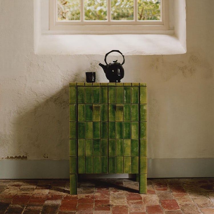A green tiled cupboard sites below a a window with a black teapot and cup on top